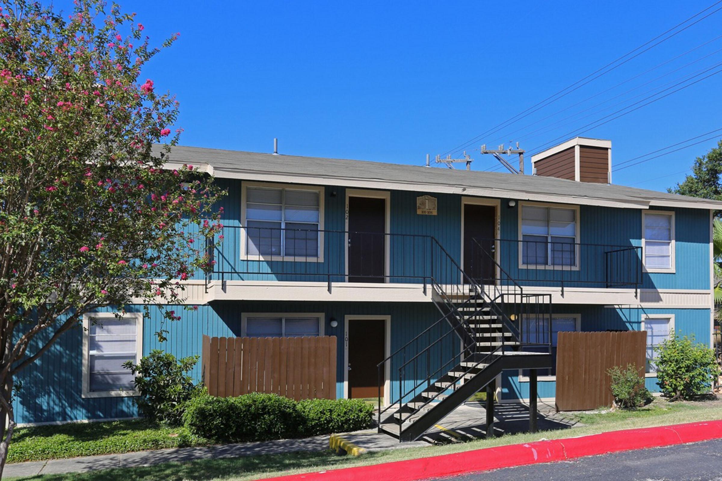 Two-story apartment building with blue and white exterior, featuring a central staircase. The building is surrounded by greenery and has a red curb in the foreground. There are visible windows and a small fence along the walkway. The sky is blue and clear.