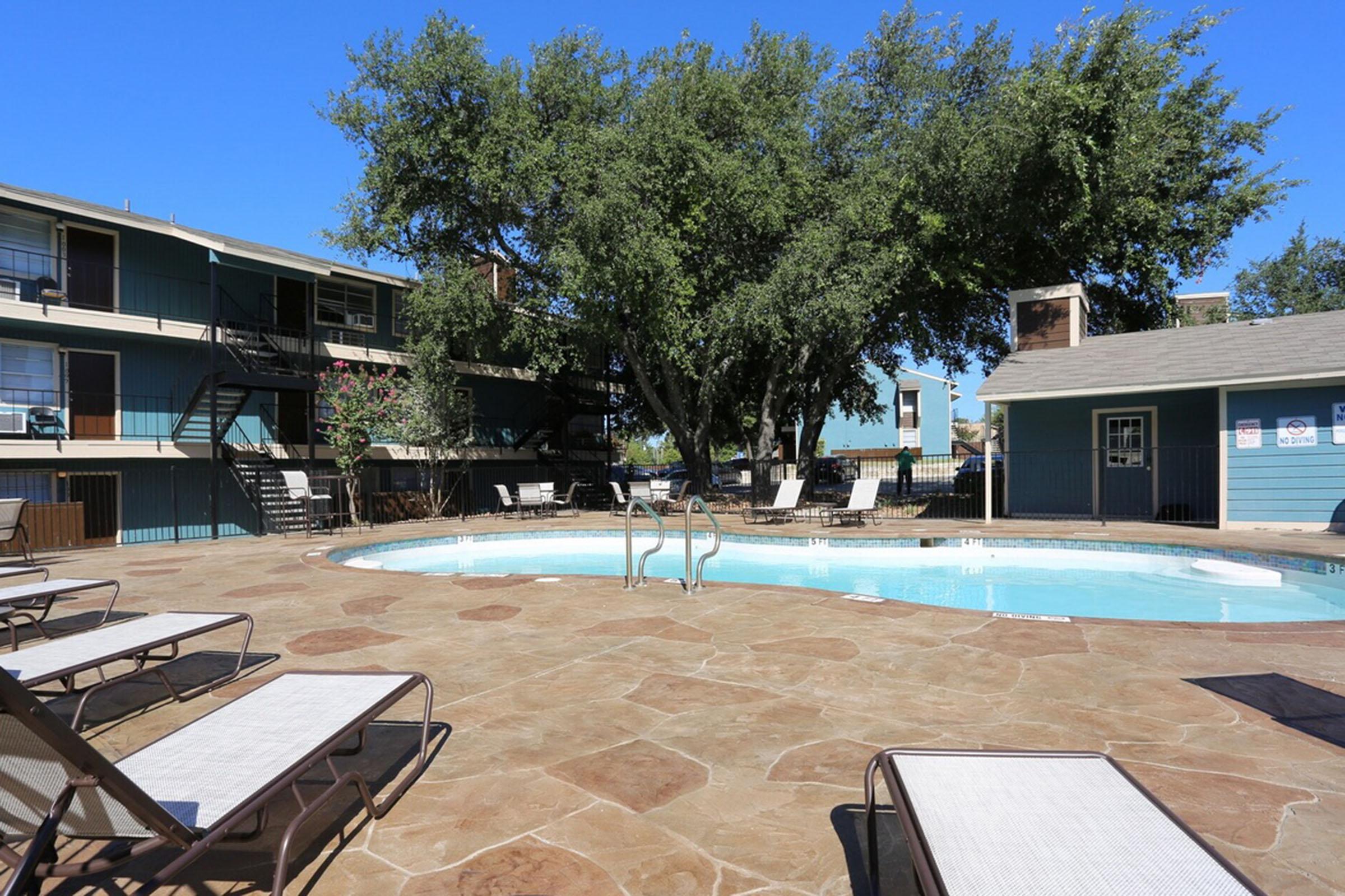 A swimming pool surrounded by lounge chairs and a stone patio. Lush green trees provide shade, and nearby, two multi-story buildings with balconies are visible. The sky is clear and blue, creating a relaxing outdoor environment.
