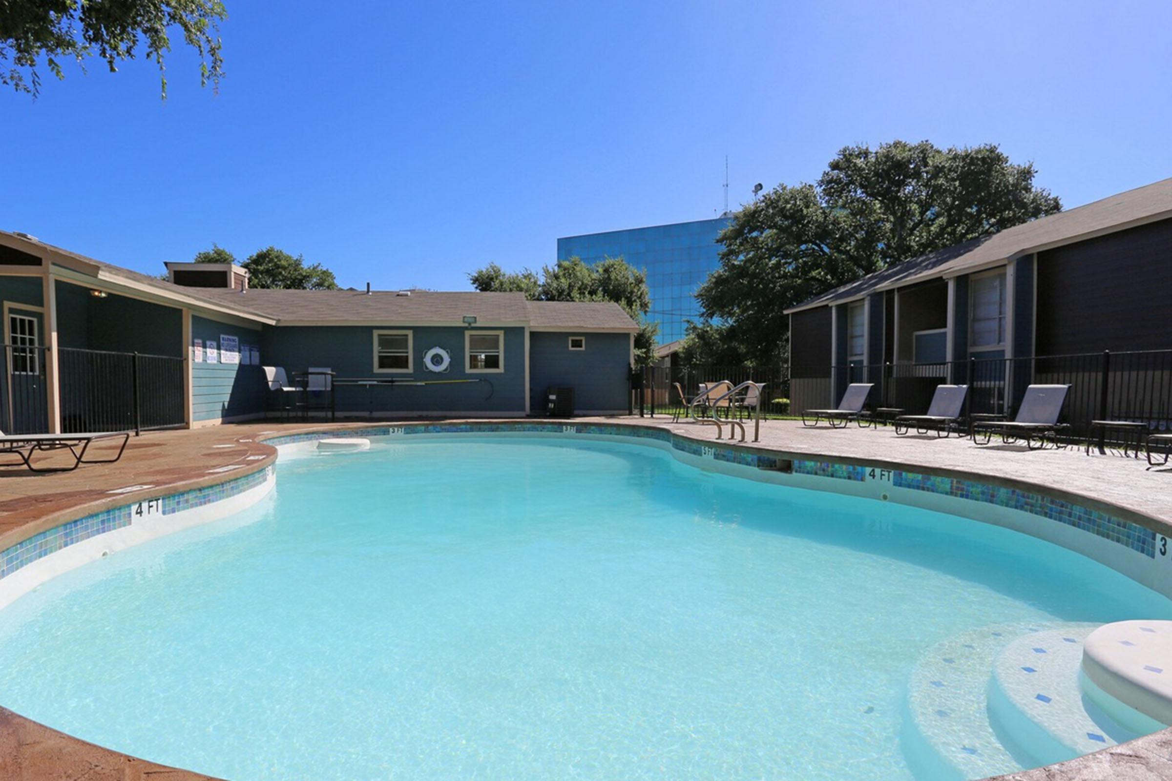 A sunny pool area featuring clear blue water, surrounded by lounge chairs. On one side, there are two buildings with dark exteriors, and in the background, a tall glass building can be seen. The sky is bright blue with a few trees near the pool area, creating a relaxing atmosphere.