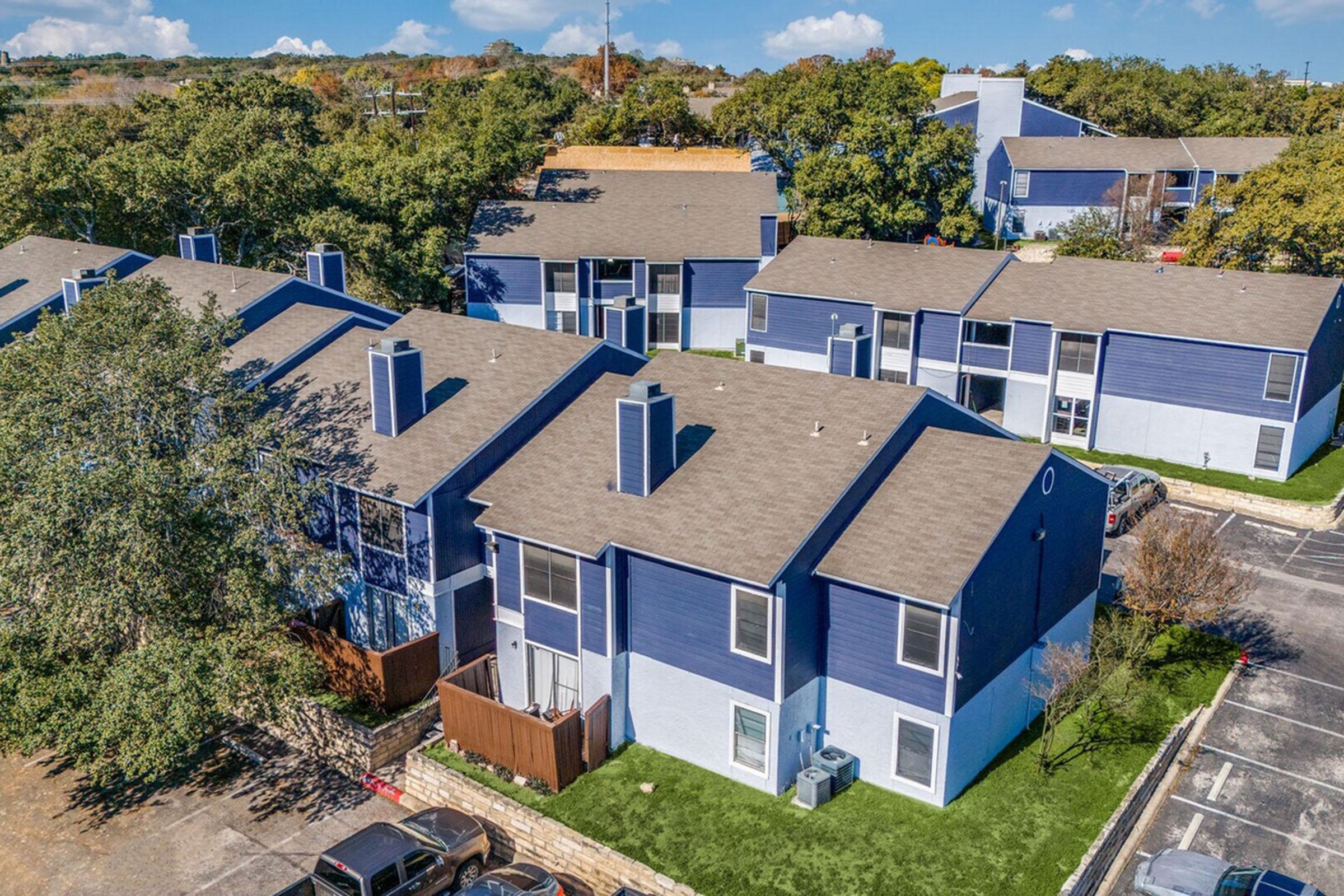 Aerial view of a residential complex featuring several two-story blue apartment buildings surrounded by trees and green lawns. Some parking spaces are visible in the foreground, with a few parked cars. The sky is clear with a few clouds.