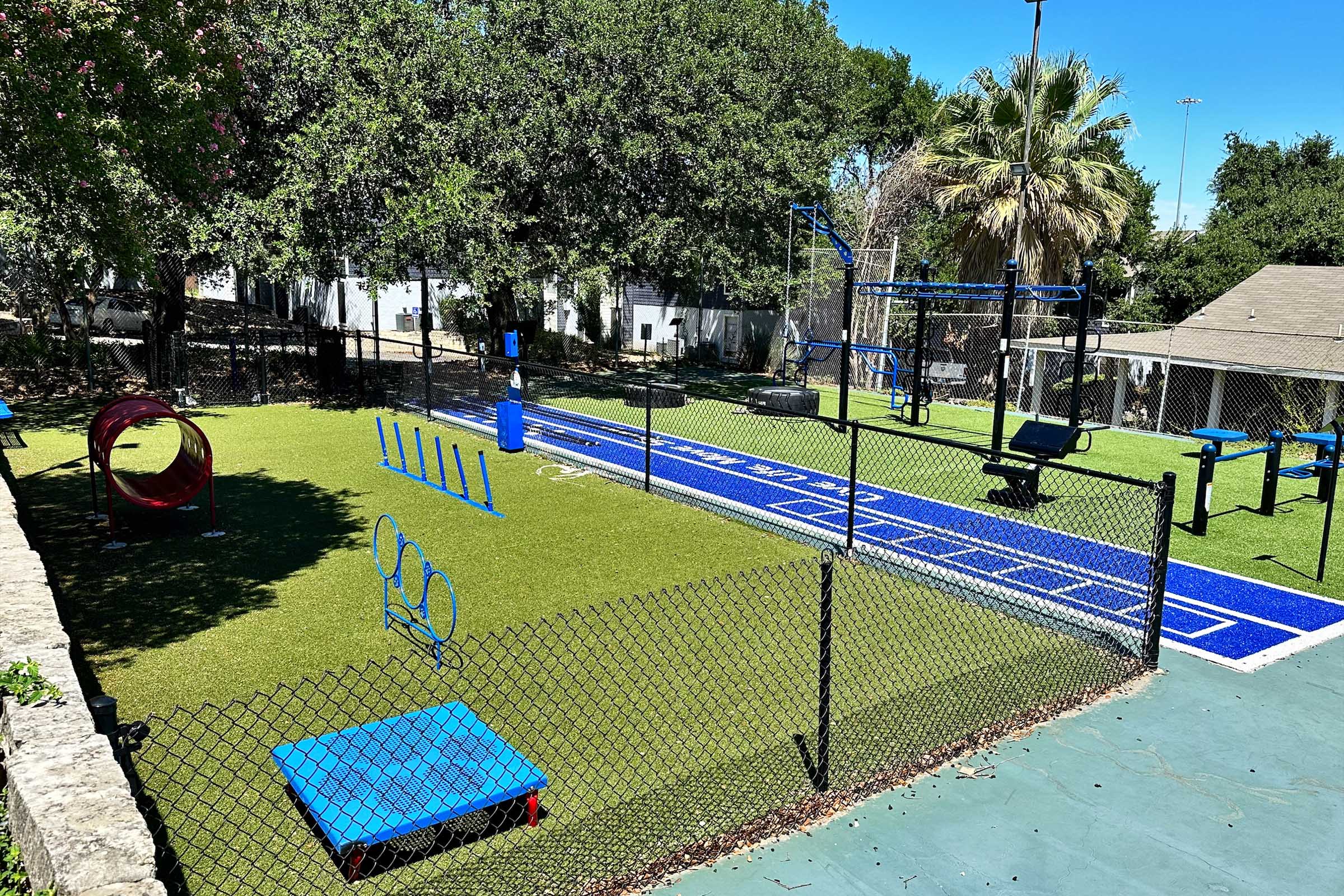 A vibrant outdoor fitness area featuring blue and green exercise equipment, agility hurdles, and a circular tunnel on artificial turf. Surrounded by a chain-link fence, the space is shaded by trees, with clear blue skies above. Ideal for exercise and training activities.