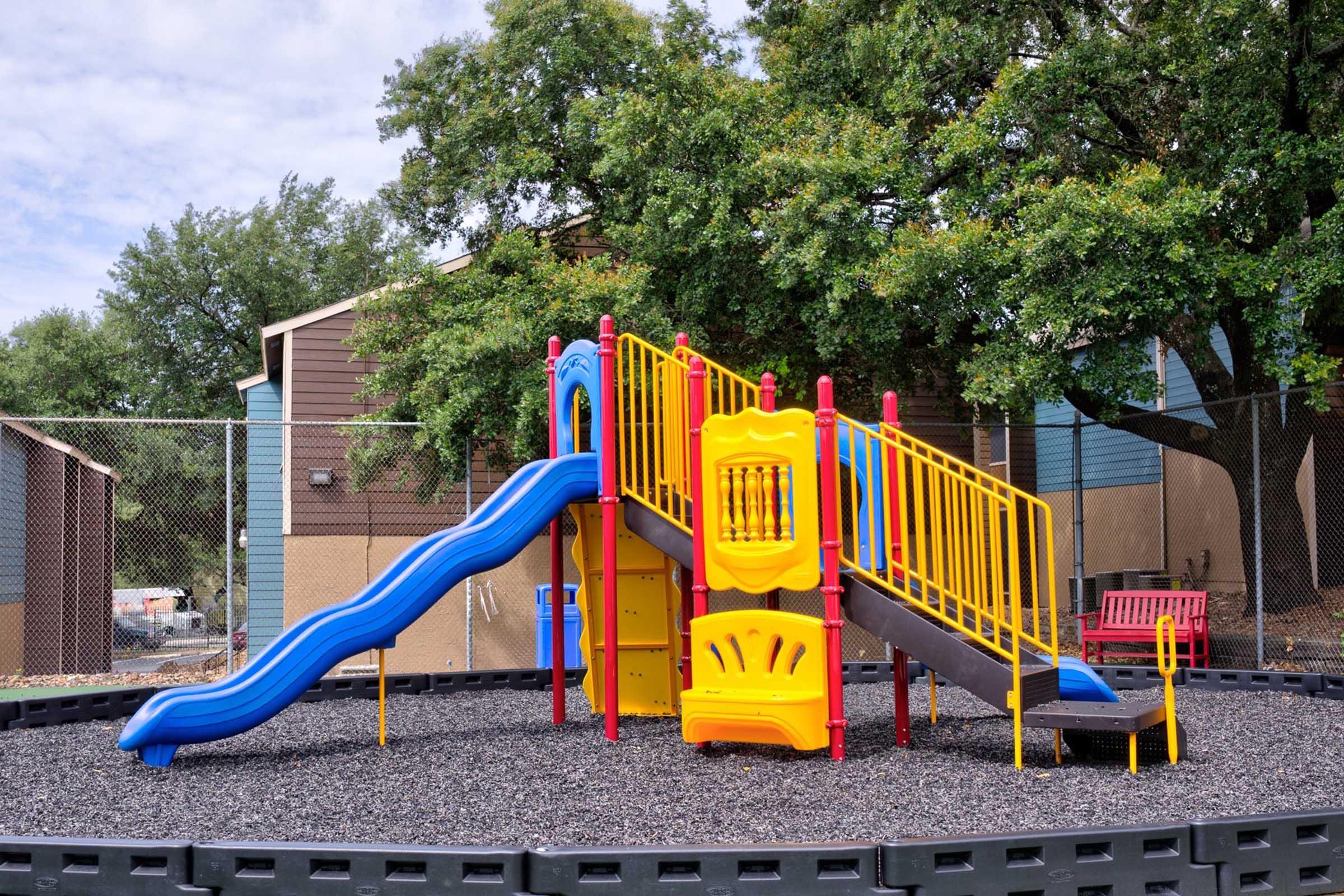 A colorful playground set featuring a blue slide, yellow climbing structure, and safety barriers, surrounded by a black rubber surface. In the background, there are trees and a building, creating a playground environment suitable for young children.