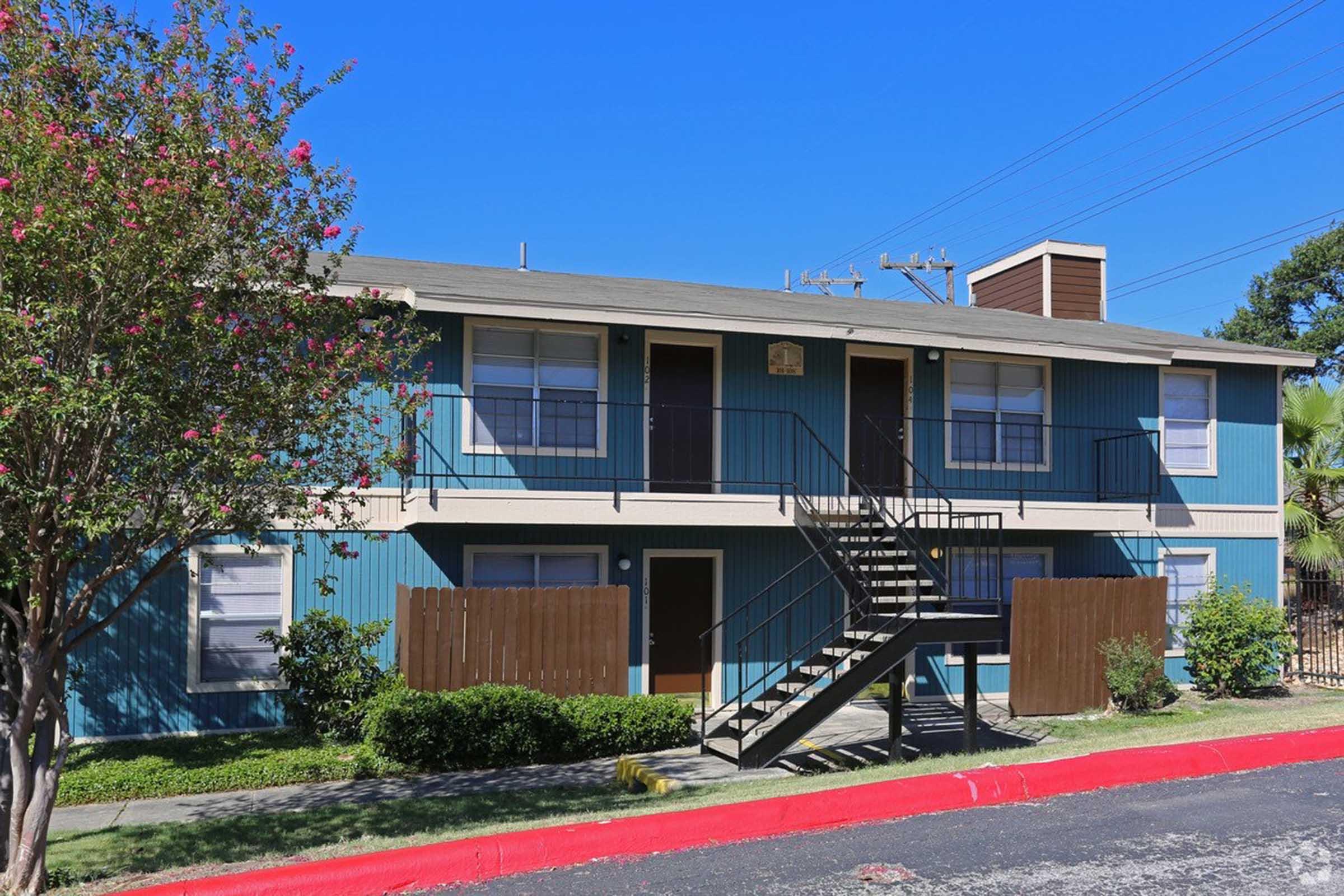 A two-story apartment building with blue and beige siding, featuring a central staircase leading to the second floor. Surrounding the building are green bushes and a small tree with pink flowers. The building has several windows and a sidewalk in front, against a clear blue sky.