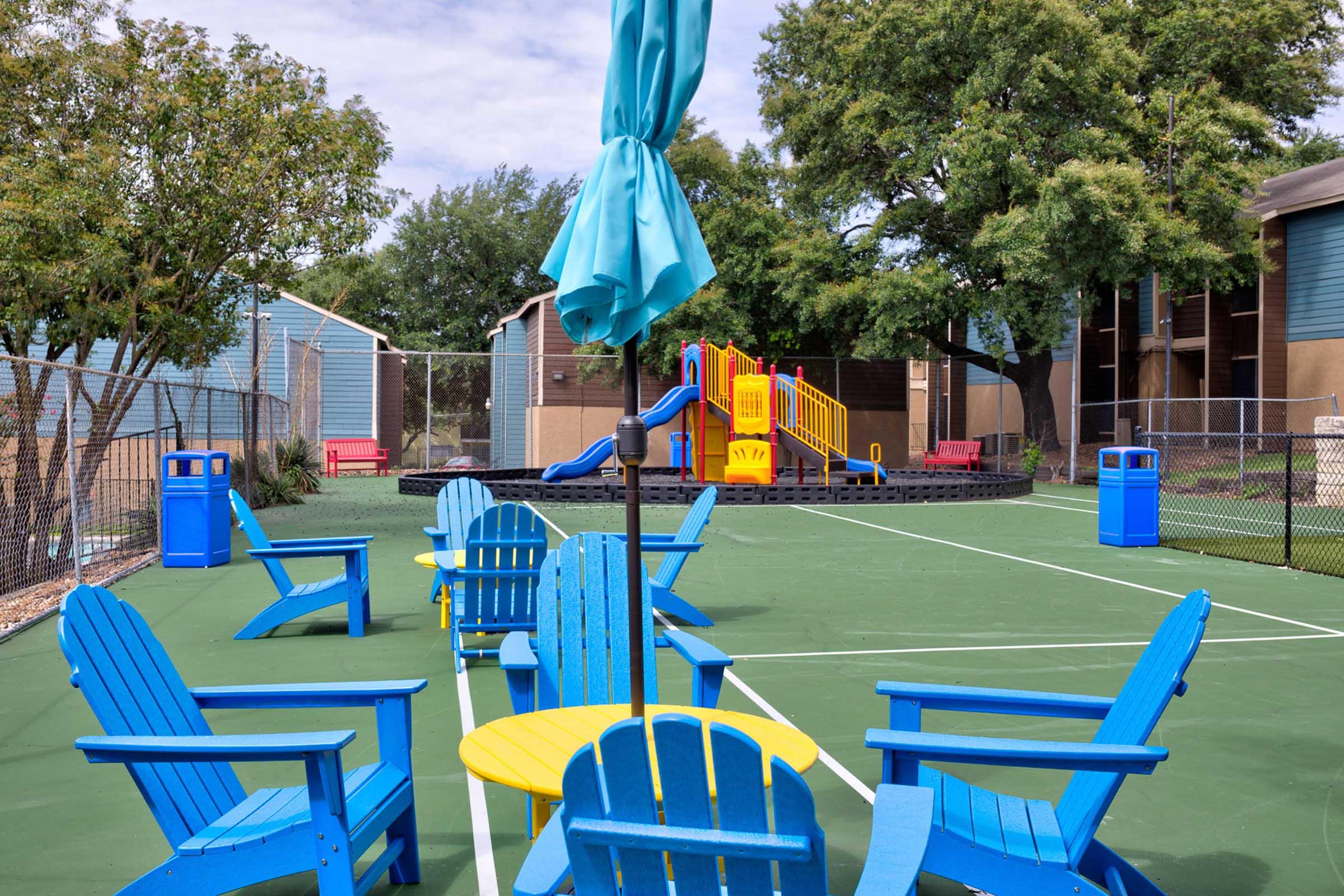Colorful blue Adirondack chairs surround a yellow table on a green playground surface. In the background, a playground with a slide and climbing structure is visible, surrounded by trees and a fence. There are also red benches and trash bins nearby, creating a welcoming recreational area.