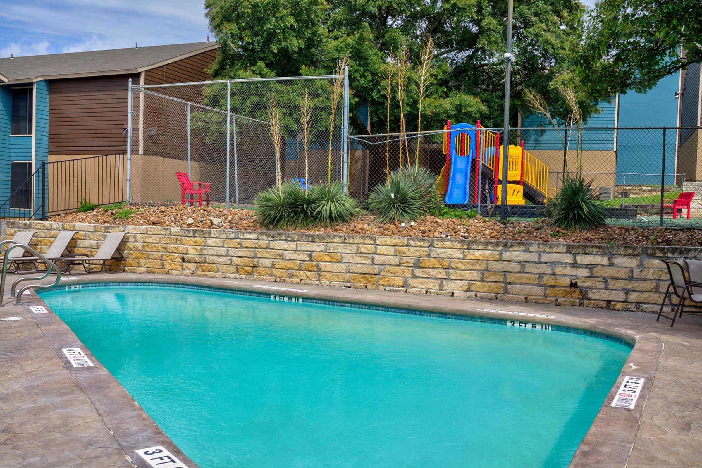 A clear blue swimming pool surrounded by a stone patio and metal fencing. Green landscaping and colorful playground equipment are visible in the background, with chairs placed around the pool area. Bright sunlight illuminates the scene, creating a welcoming atmosphere for leisure and recreation.