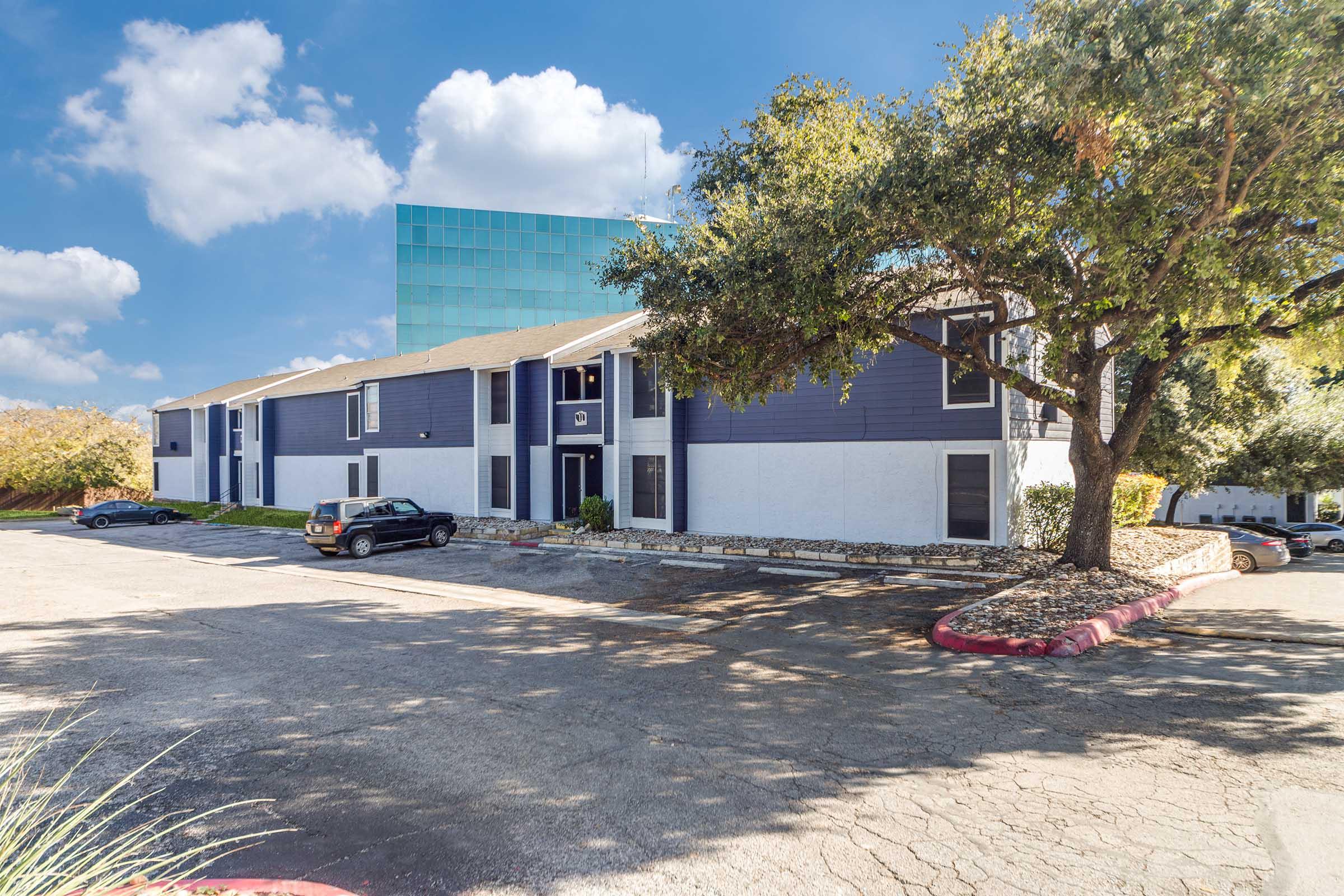 A multi-story apartment building with a blue and white exterior, surrounded by trees. A parking lot in the foreground has several parked cars. In the background, a large glass building reflects the sky, contributing to a sunny, clear day atmosphere.