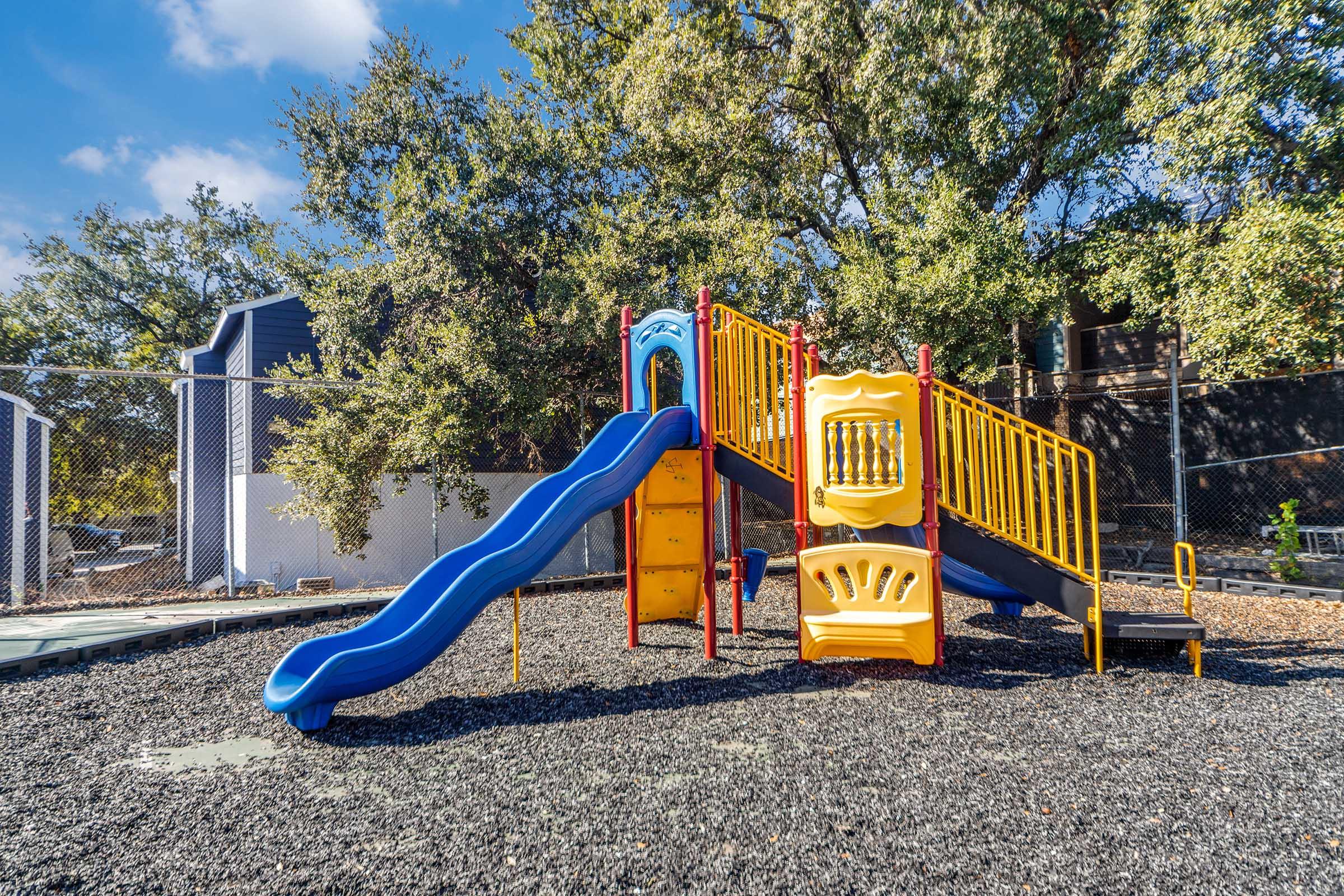 Colorful playground equipment featuring a blue slide and play structures in yellow and red. Surrounding the area is a layer of black rubber mulch, and trees provide shade in the background. A chain-link fence encloses the playground.