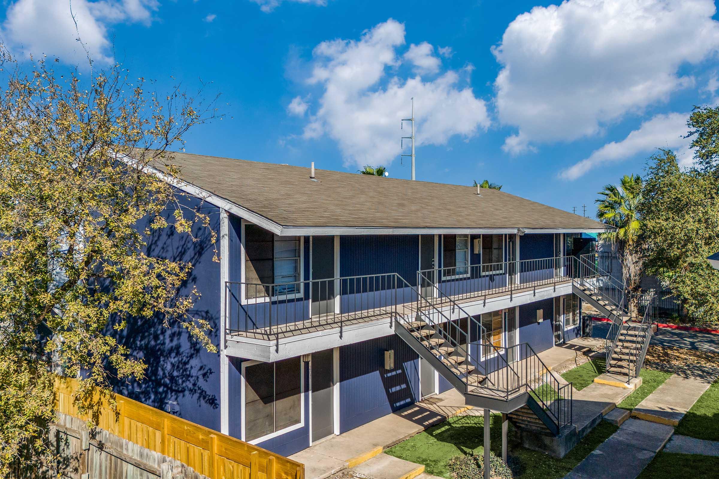 Two-story apartment building with blue walls, featuring a series of stairs leading to individual units. Surrounding greenery includes trees and palm trees, under a bright blue sky with scattered clouds. A pathway runs in front of the building, providing access to the entrances.