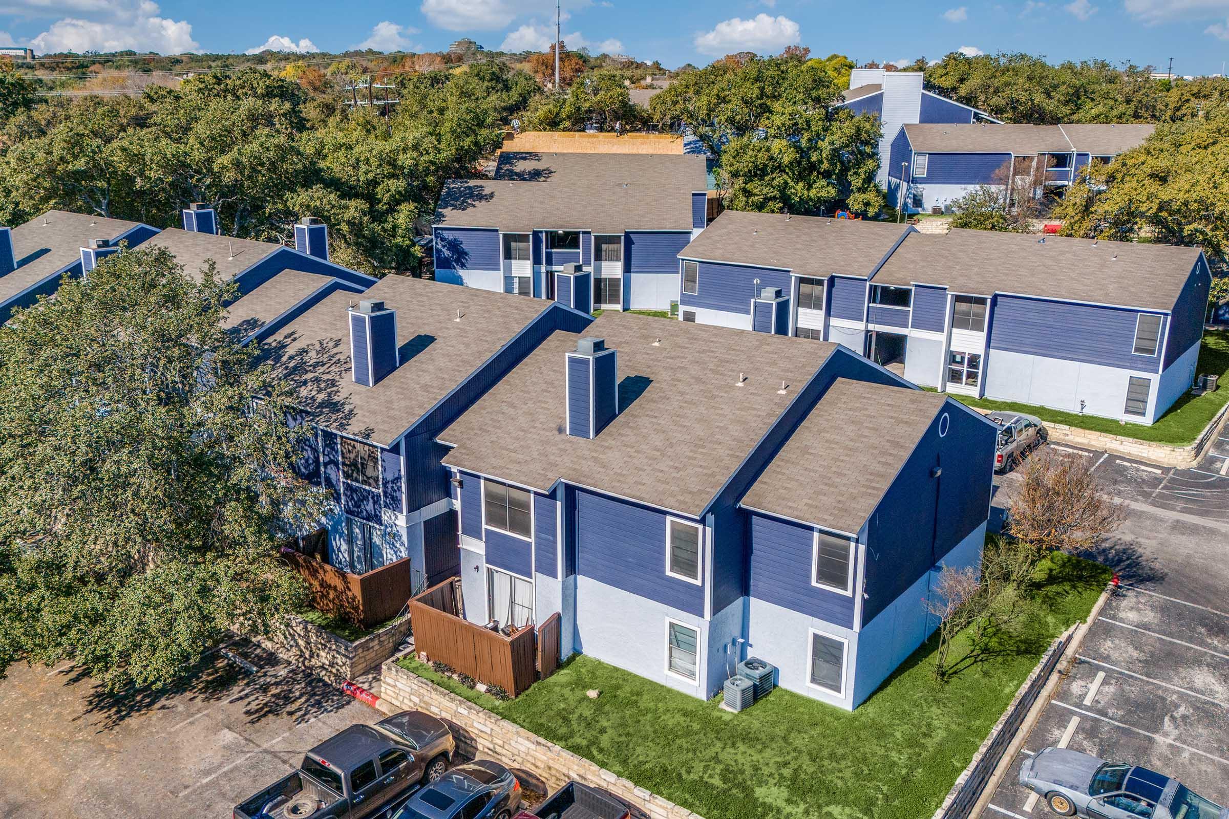 Aerial view of a residential complex featuring two-story blue apartment buildings with tan roofs surrounded by green trees and well-maintained lawns. Parking spaces are visible in the foreground, and a clear blue sky with a few clouds is seen above.