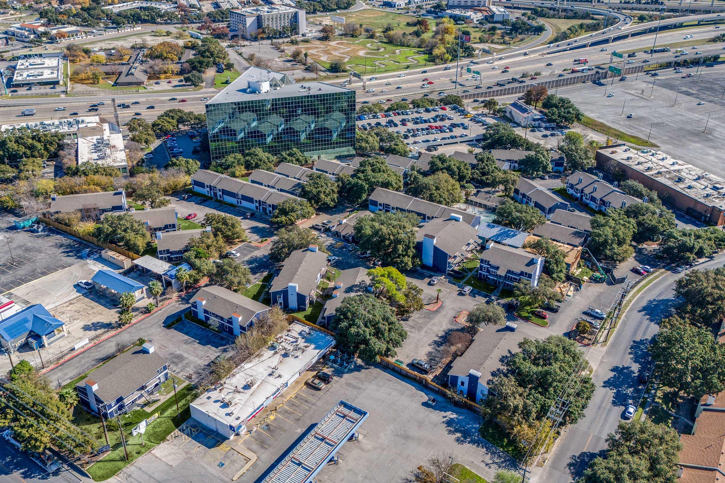 Aerial view of a mixed-use development featuring multiple low-rise buildings, parking lots, and a nearby highway. The scene includes green landscaping with trees, a large glass building in the background, and various parked cars, showcasing a blend of residential and commercial areas.