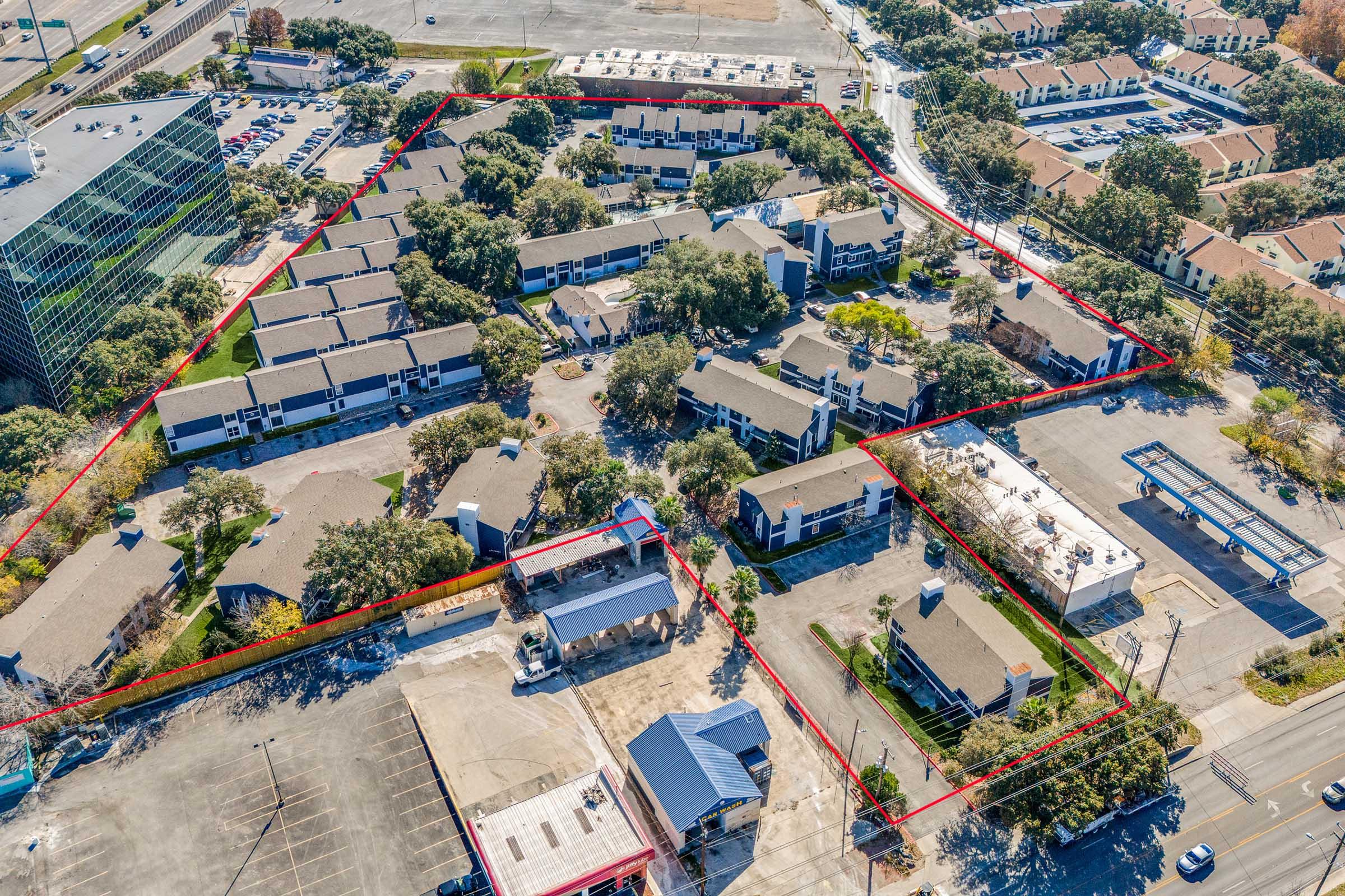 Aerial view of a complex featuring several buildings surrounded by greenery, outlined in red. The area includes parking lots and open spaces, with some structures appearing to be residential or commercial. Nearby, streets and additional properties are visible, showcasing urban development.