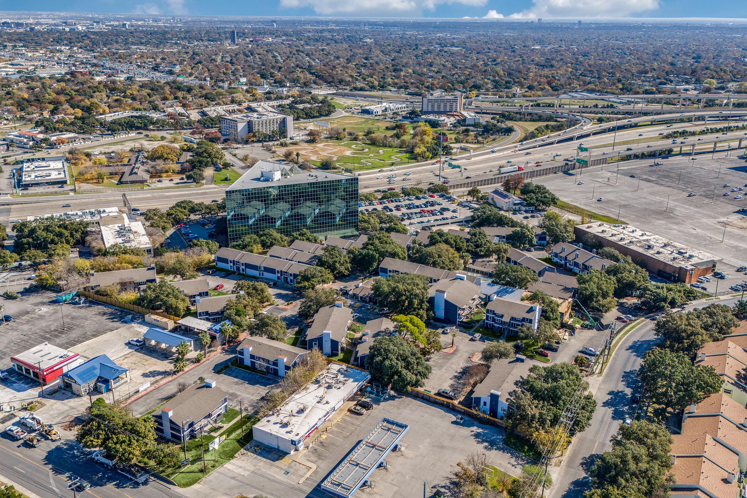 Aerial view of a suburban area featuring a mix of commercial buildings and residential complexes. The landscape includes green spaces, parking lots, and intersecting highways. The scene is characterized by scattered trees and a clear blue sky, with city structures extending into the distance.