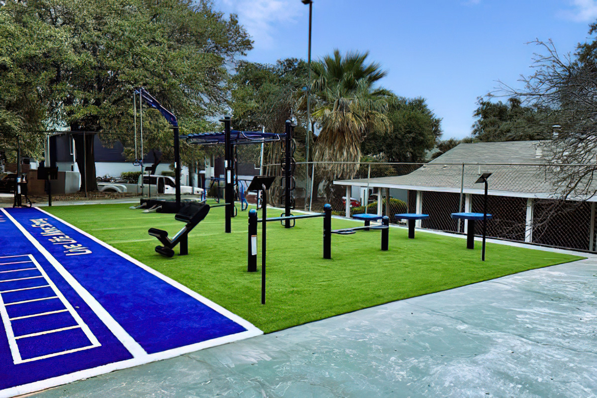 A modern outdoor gym area featuring an artificial turf surface. It includes various exercise equipment such as swings, pull-up bars, and other fitness tools, all set against a backdrop of trees and a residential building under a clear blue sky.