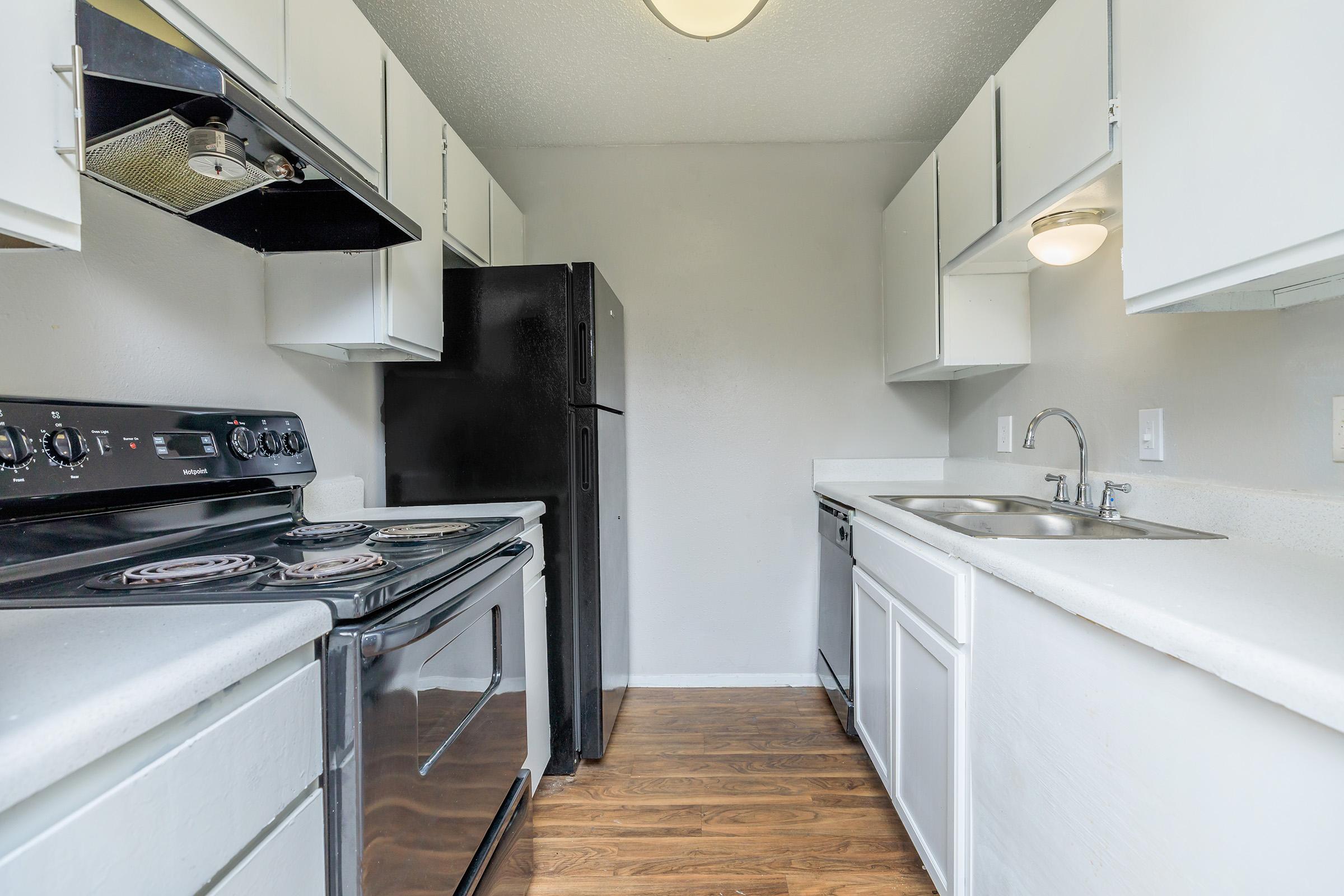 Modern kitchen featuring white cabinets, a black refrigerator, a black stove with an oven, and a stainless steel sink with a single faucet. The countertops are light-colored, and the flooring is a warm wood tone. The walls are painted a neutral color, providing a bright and open atmosphere.