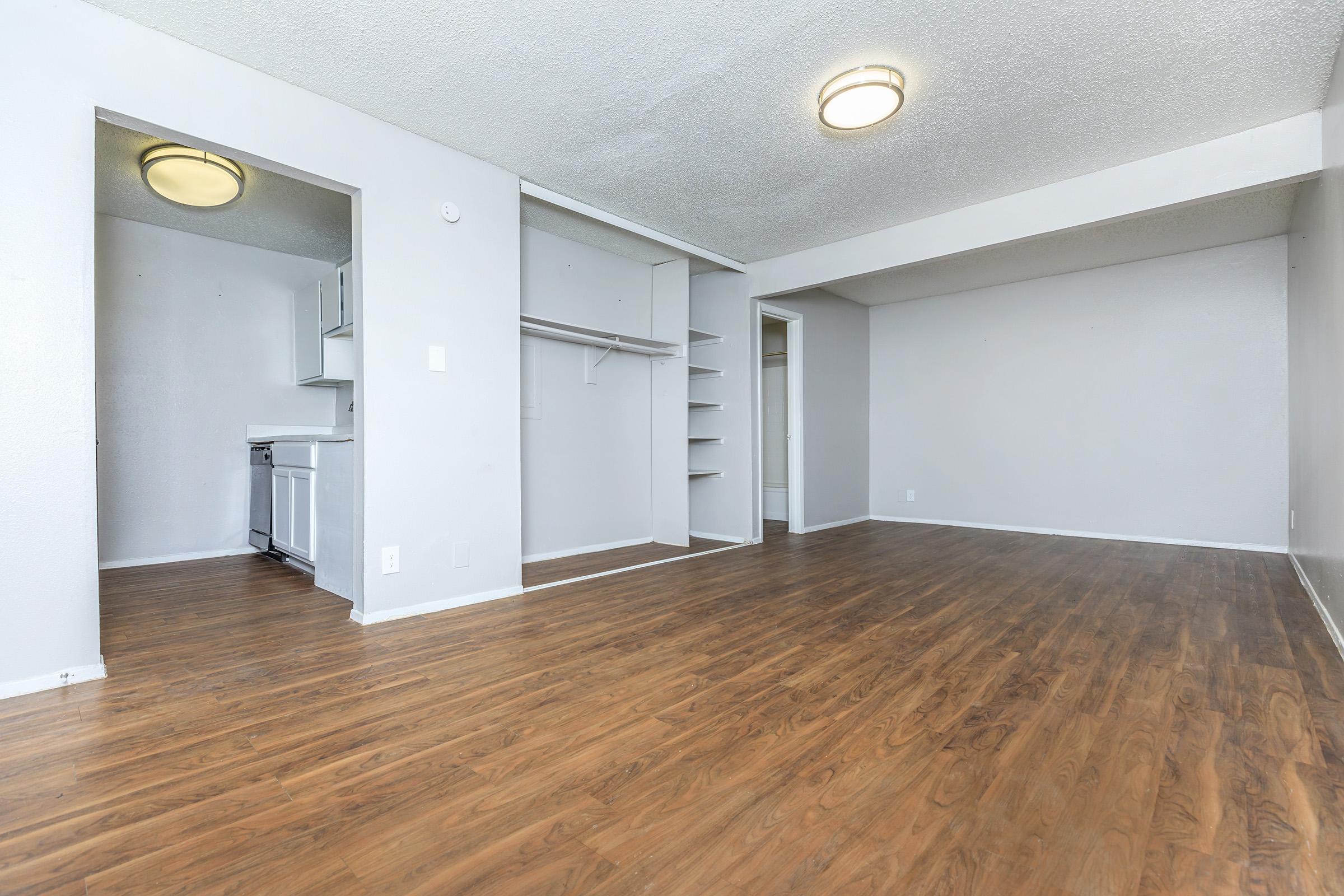 Interior view of a spacious, empty room featuring light gray walls and wood laminate flooring. On the left, there is a small kitchen area visible, with a white fridge and cabinetry. The room has a large window, ample natural light, and a simple ceiling light fixture. Closets and shelving are present along one wall.