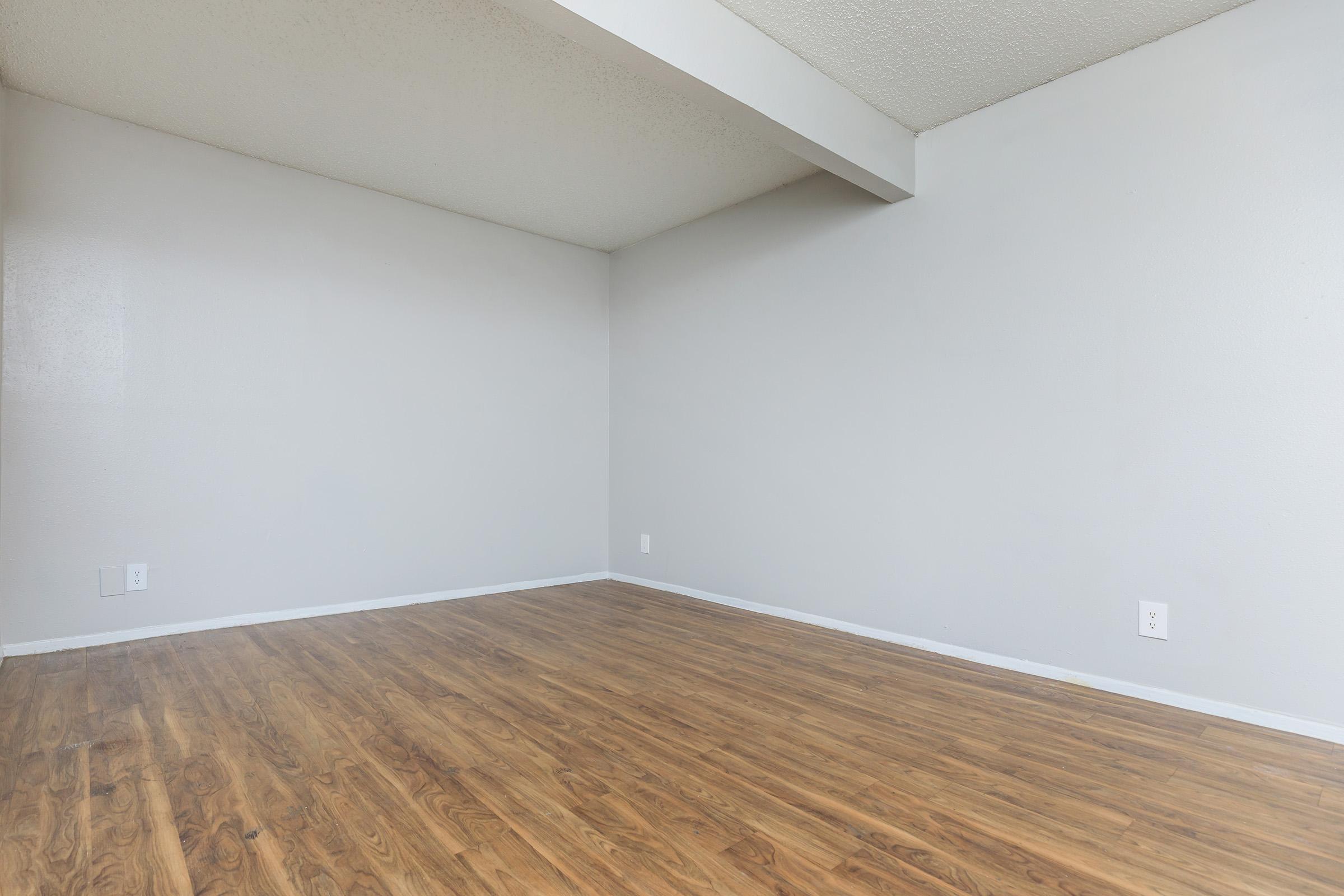 An empty room with light gray walls and a wooden floor. The space features a single beam across the ceiling and minimal decor, creating a simple and spacious atmosphere. Natural light is absent, giving the room a neutral appearance.