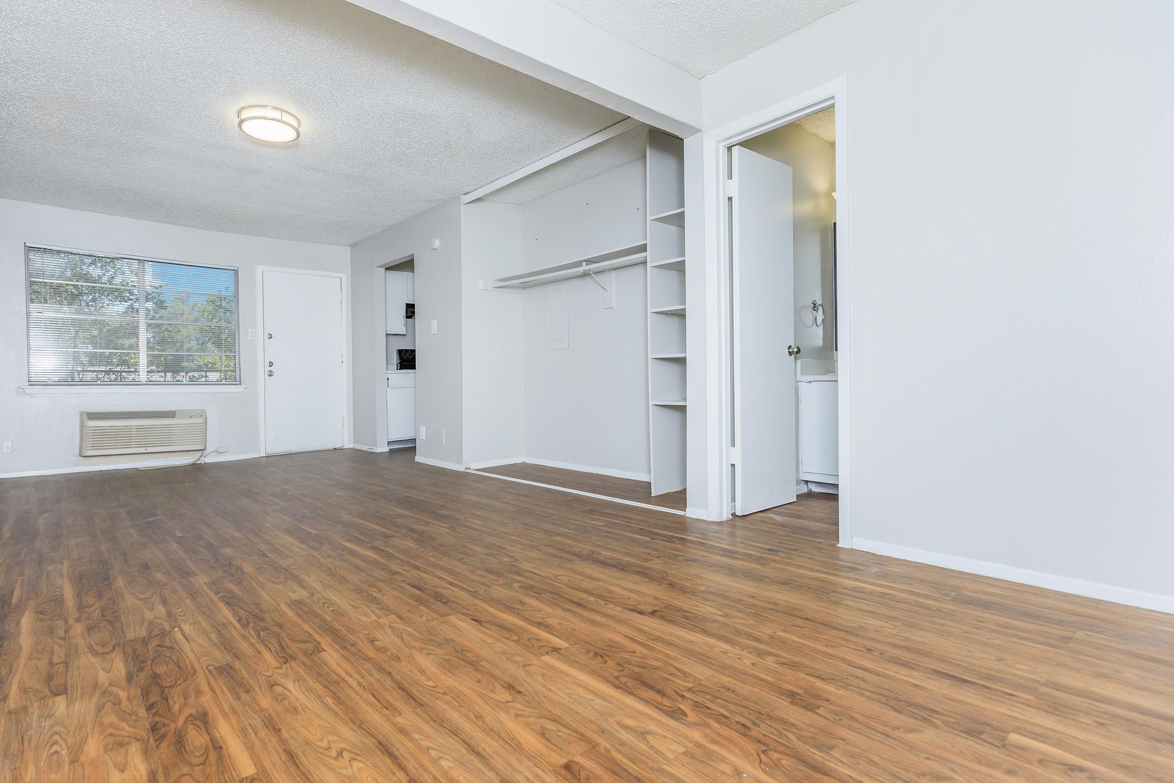 Bright and spacious interior of an apartment showcasing a living area with laminate flooring, a closet space, and a doorway leading to a bathroom. A window allows natural light to enter, and the background hints at a kitchen area. The walls are painted a light color, creating a clean and modern look.