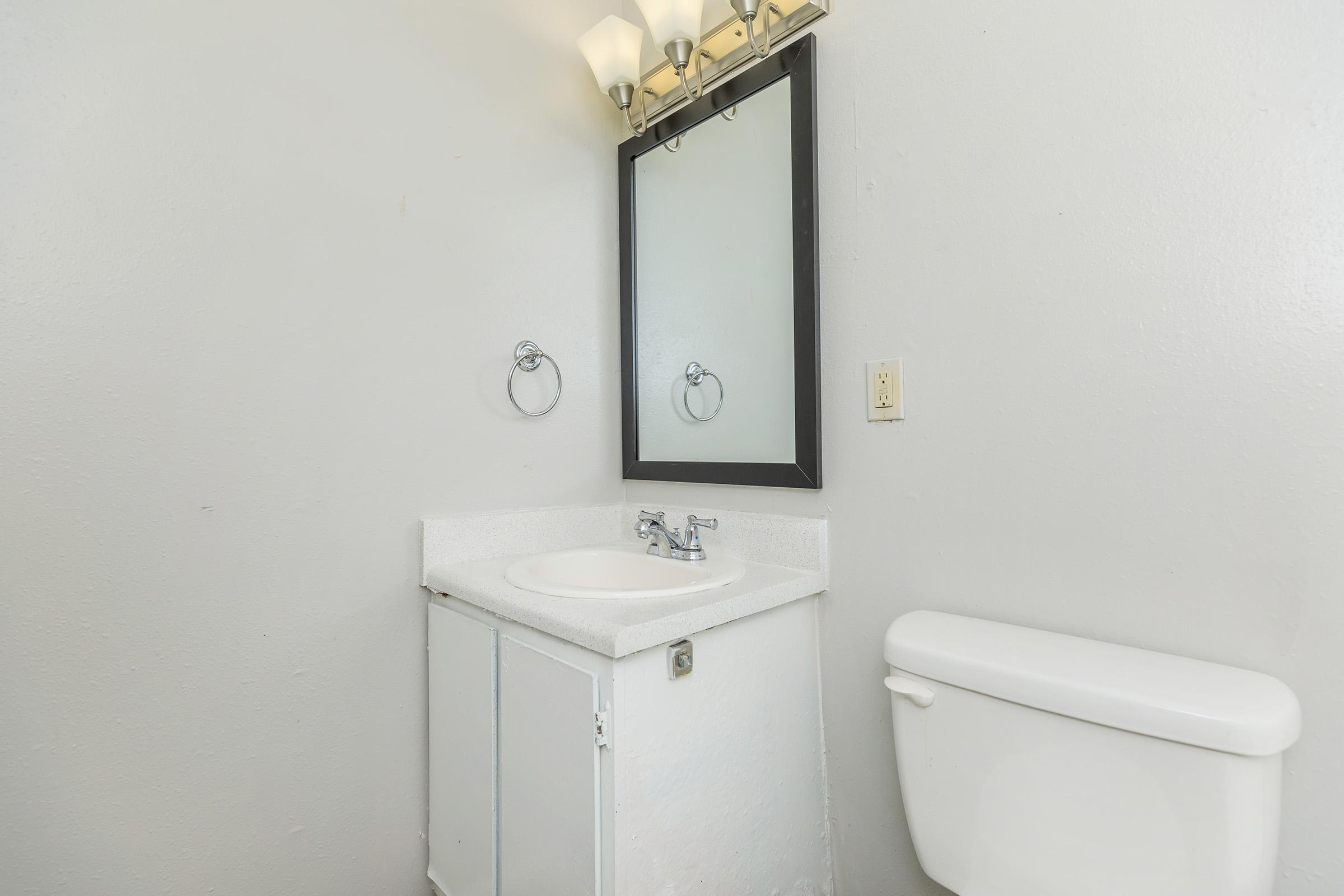 A small bathroom featuring a white vanity with a sink, a square mirror above it, and wall-mounted light fixtures. There are hand towel rings on either side of the mirror, and a white toilet is positioned adjacent to the vanity. The walls are painted a light color, enhancing the bright ambiance.