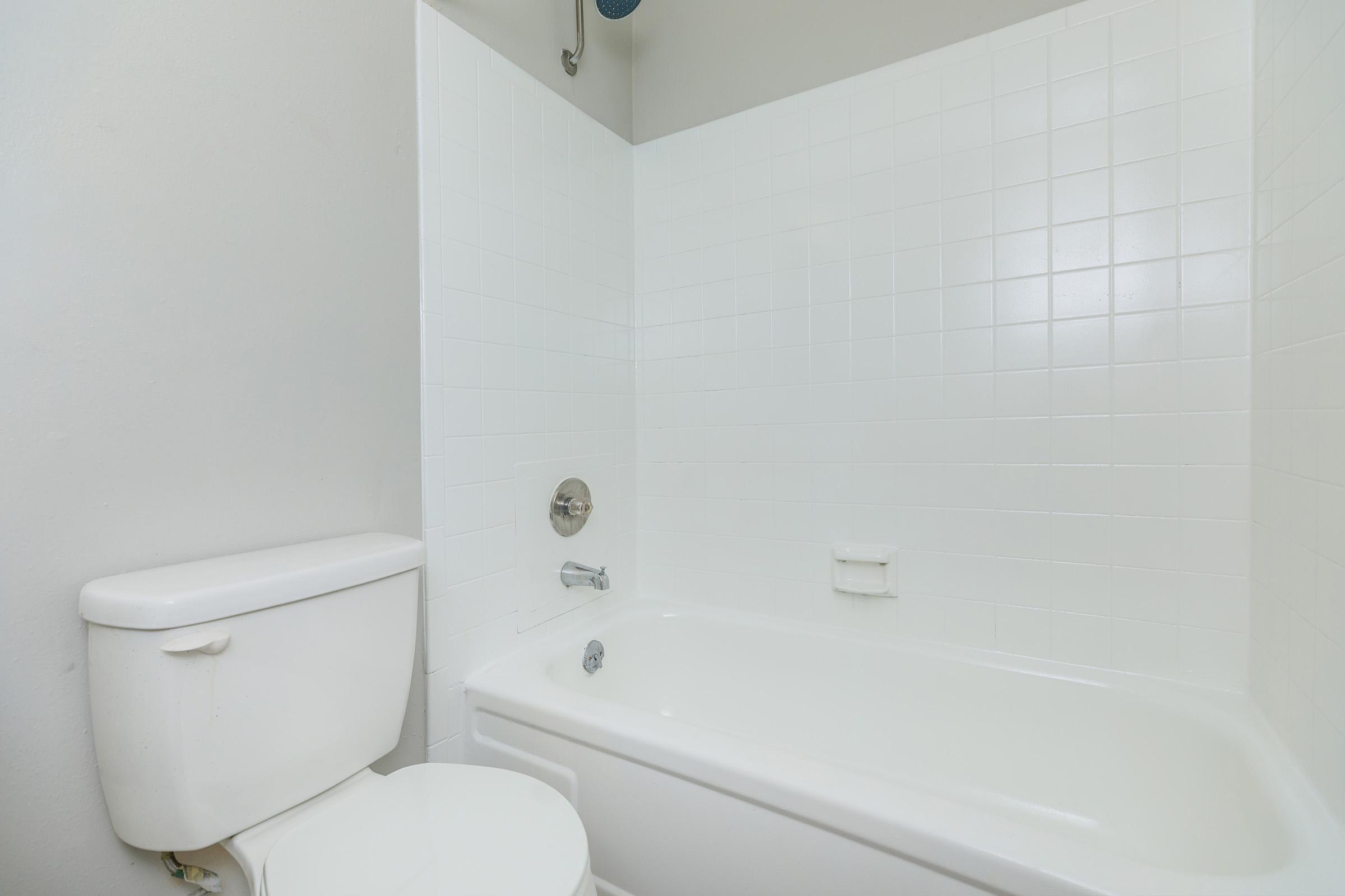 A clean, simple bathroom featuring a white bathtub with a shower, white tiled walls, a chrome faucet, and a toilet. The overall aesthetic is modern and minimalistic with neutral tones.