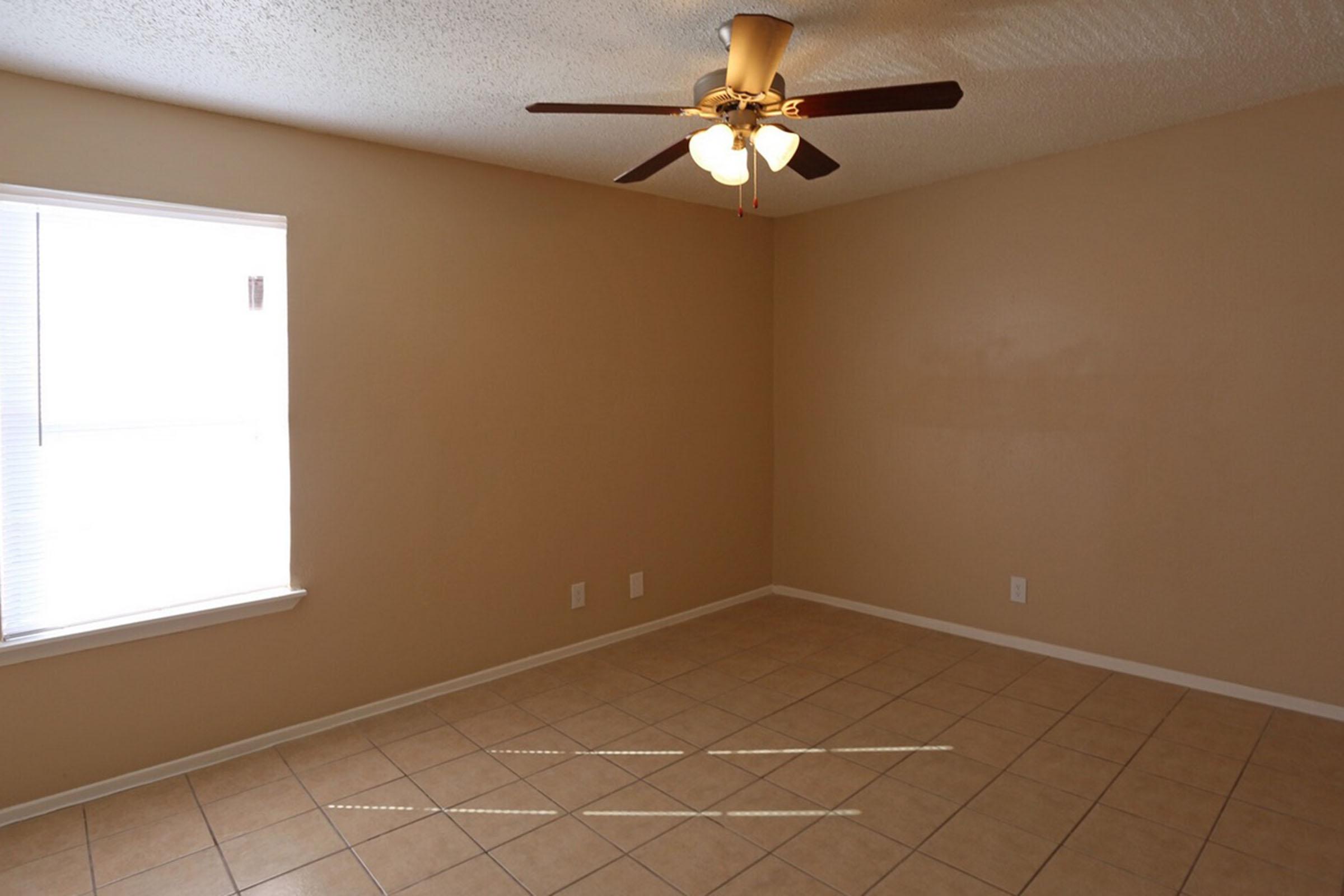 A vacant room featuring beige walls and tiled flooring. A ceiling fan with three light bulbs is mounted in the center. Natural light filters in through a window with blinds on the left side. The room is minimalistic and unadorned, suggesting it is ready for furnishing.
