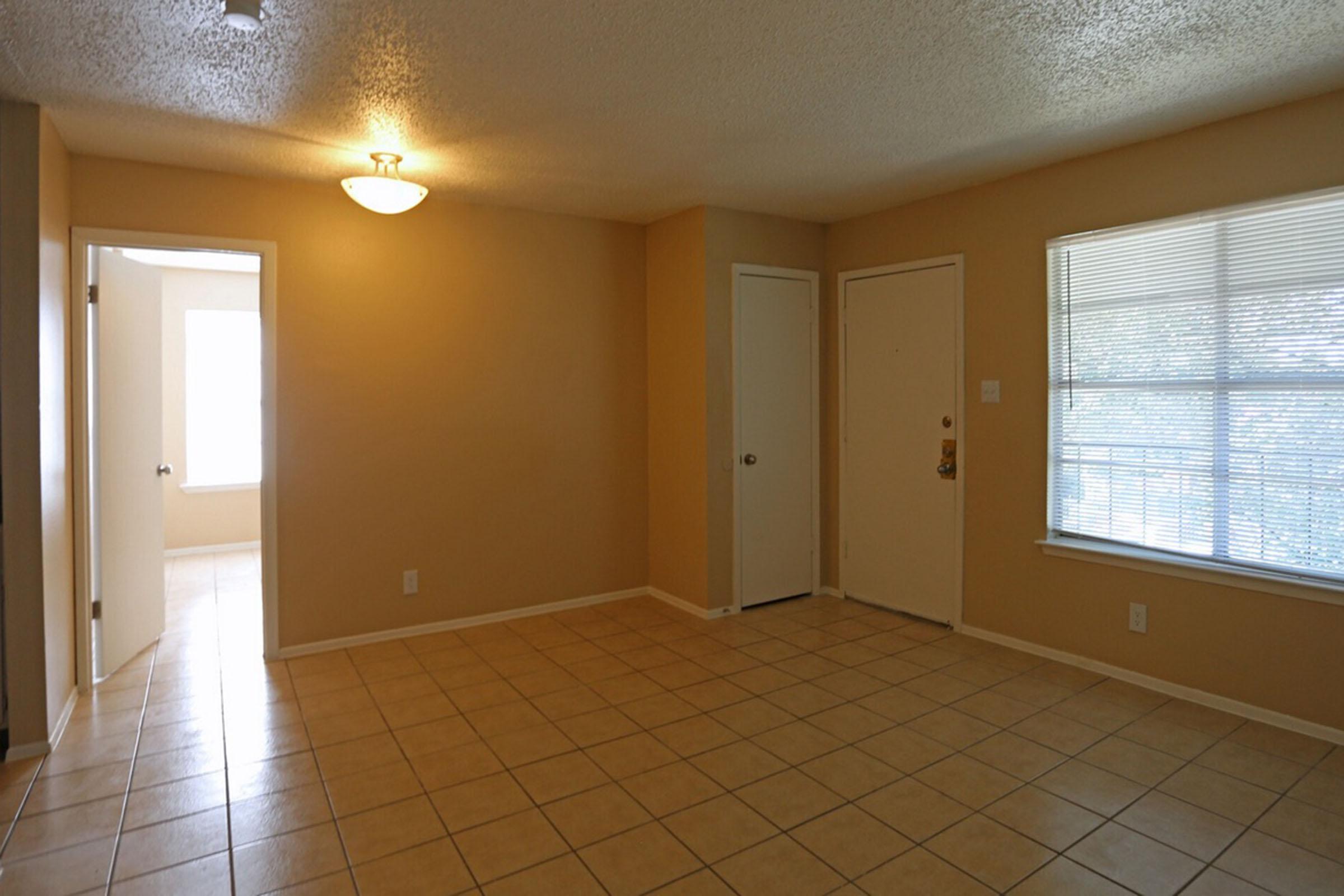 A spacious, empty room with beige walls and tile flooring. A ceiling light illuminates the area. There are two doors on the left leading to adjacent spaces, and a large window with blinds on the right, allowing natural light to enter. The overall atmosphere is bright and inviting.