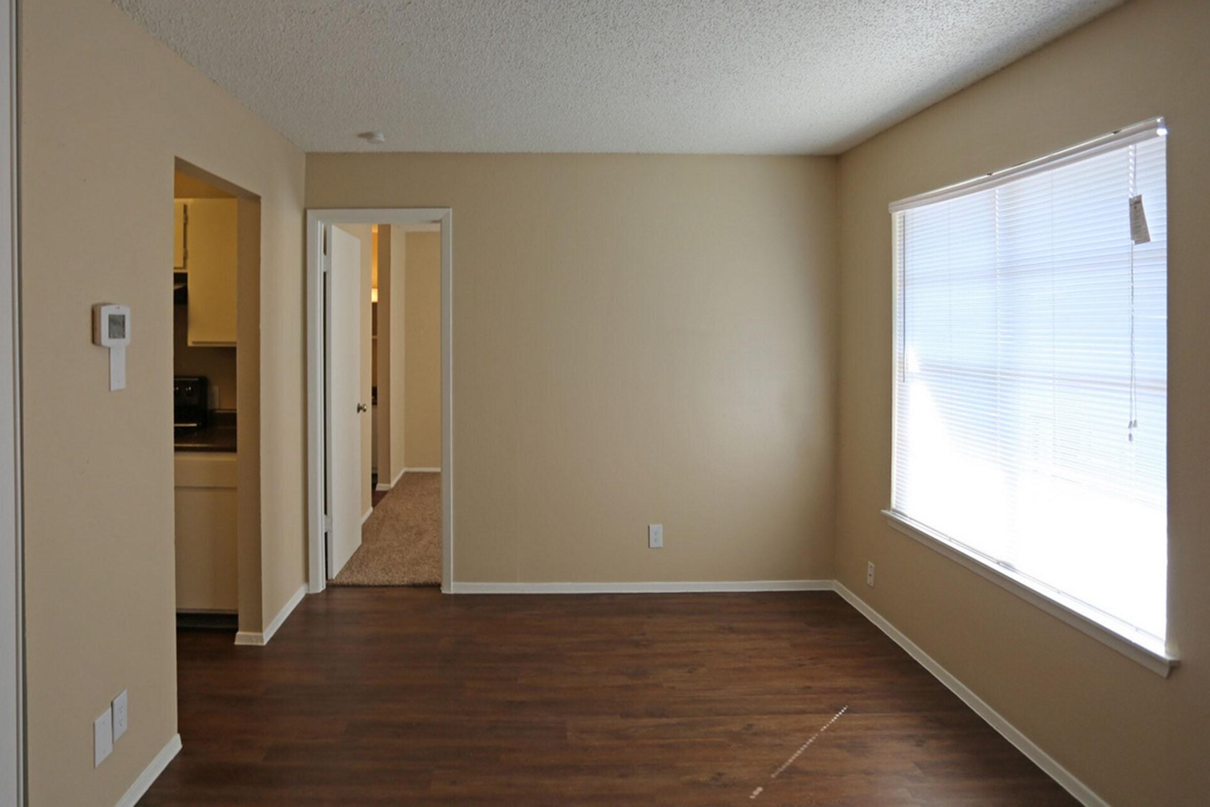A bright, empty room with light brown walls and wooden flooring. On one side, there is a large window with blinds, allowing natural light to fill the space. A doorway leads to another room, and a second door is visible on the left side, suggesting the entrance to a kitchen area.