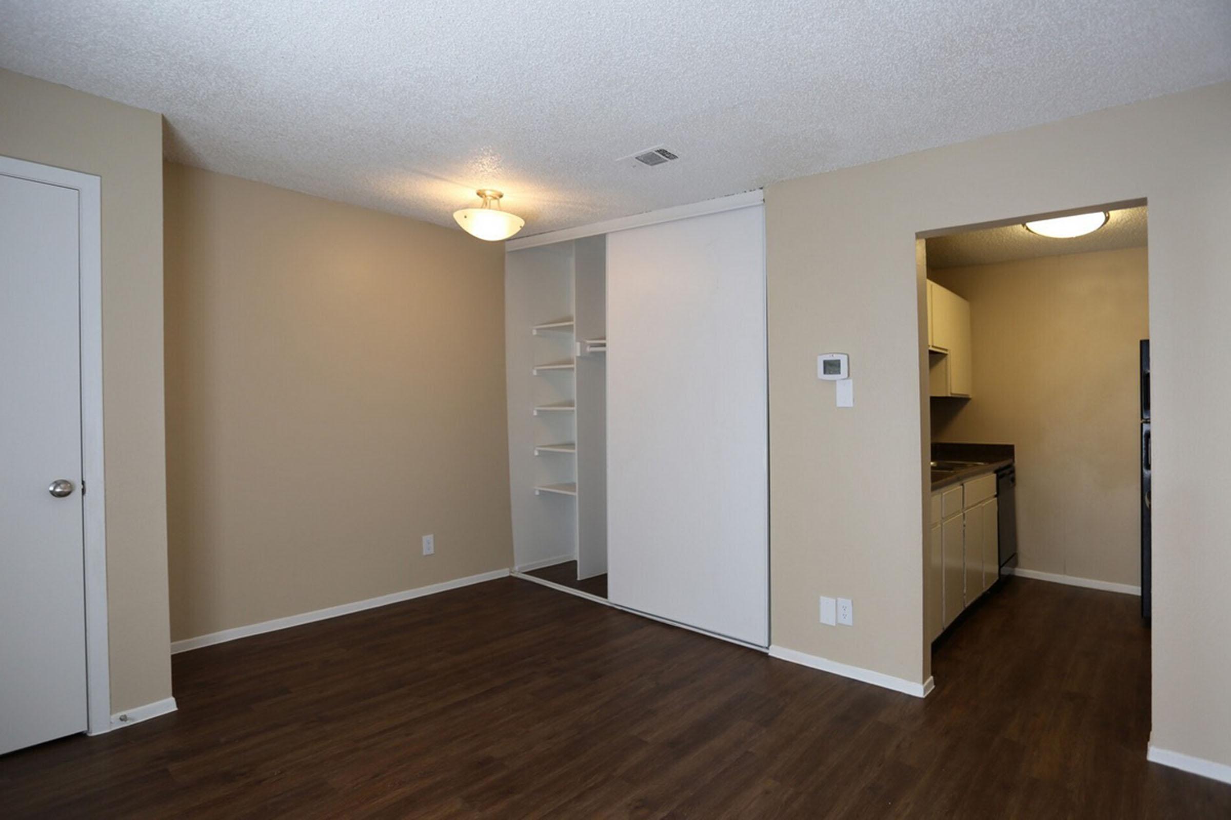 A well-lit interior of a small apartment showing a living space with light brown walls and a ceiling light. To the left, there's a sliding white closet door, and to the right is an open kitchen area with beige cabinets. The floor is wooden, contributing to a modern, minimalistic aesthetic.