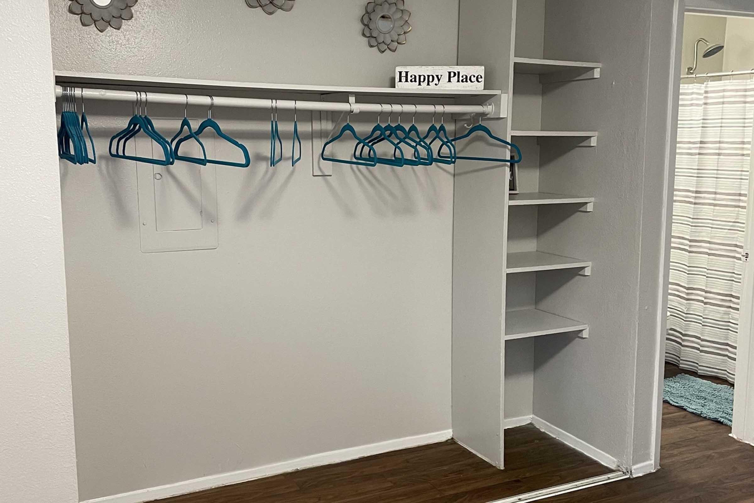 An empty closet with a white wall, featuring a hanging rod with several blue plastic hangers. There is a sign that reads "Happy Place" above the hangers. To the right, there are empty shelves and a partially visible bathroom with a shower curtain in the background. The floor is wooden.