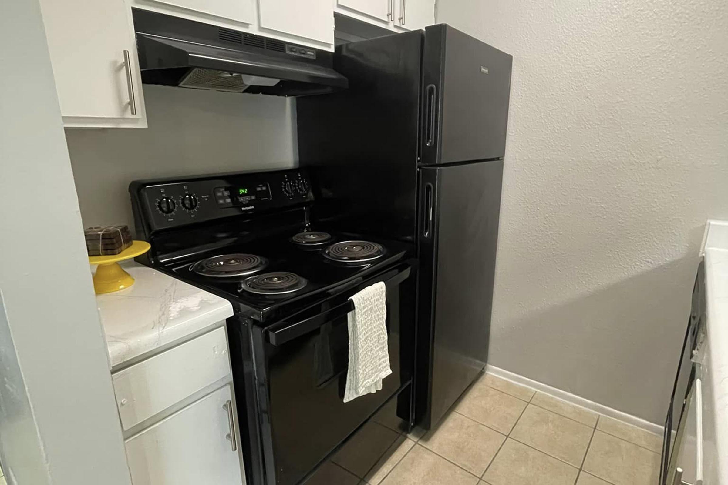 A modern kitchen featuring a black stove with four burners beneath a range hood, a black refrigerator, and light-colored cabinets. The stove has a white dish towel hanging from it, and the tiled floor adds a clean, contemporary look to the space.