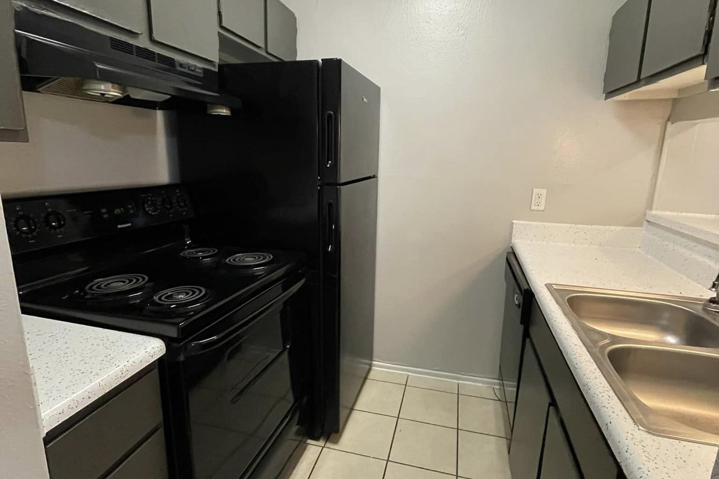 A compact kitchen featuring black appliances, including a stove and refrigerator, with light-colored countertops and dark cabinets. The space has a tiled floor and is well-lit, creating a functional and modern cooking area.