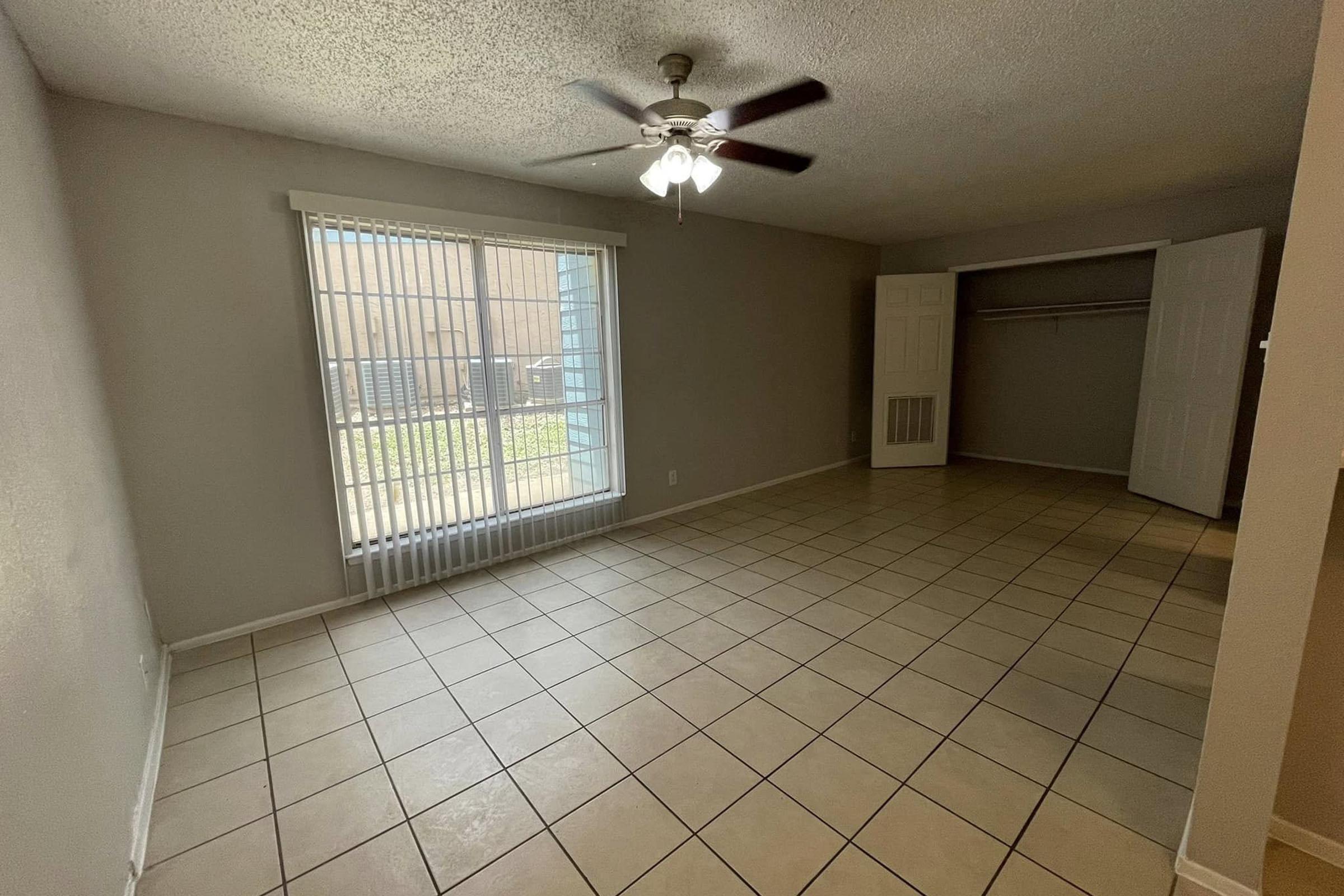 Spacious, empty room featuring tiled flooring, a ceiling fan, and large windows with blinds allowing natural light. An adjacent closet with sliding doors is visible. The walls are painted in a neutral color, creating a bright and inviting atmosphere.