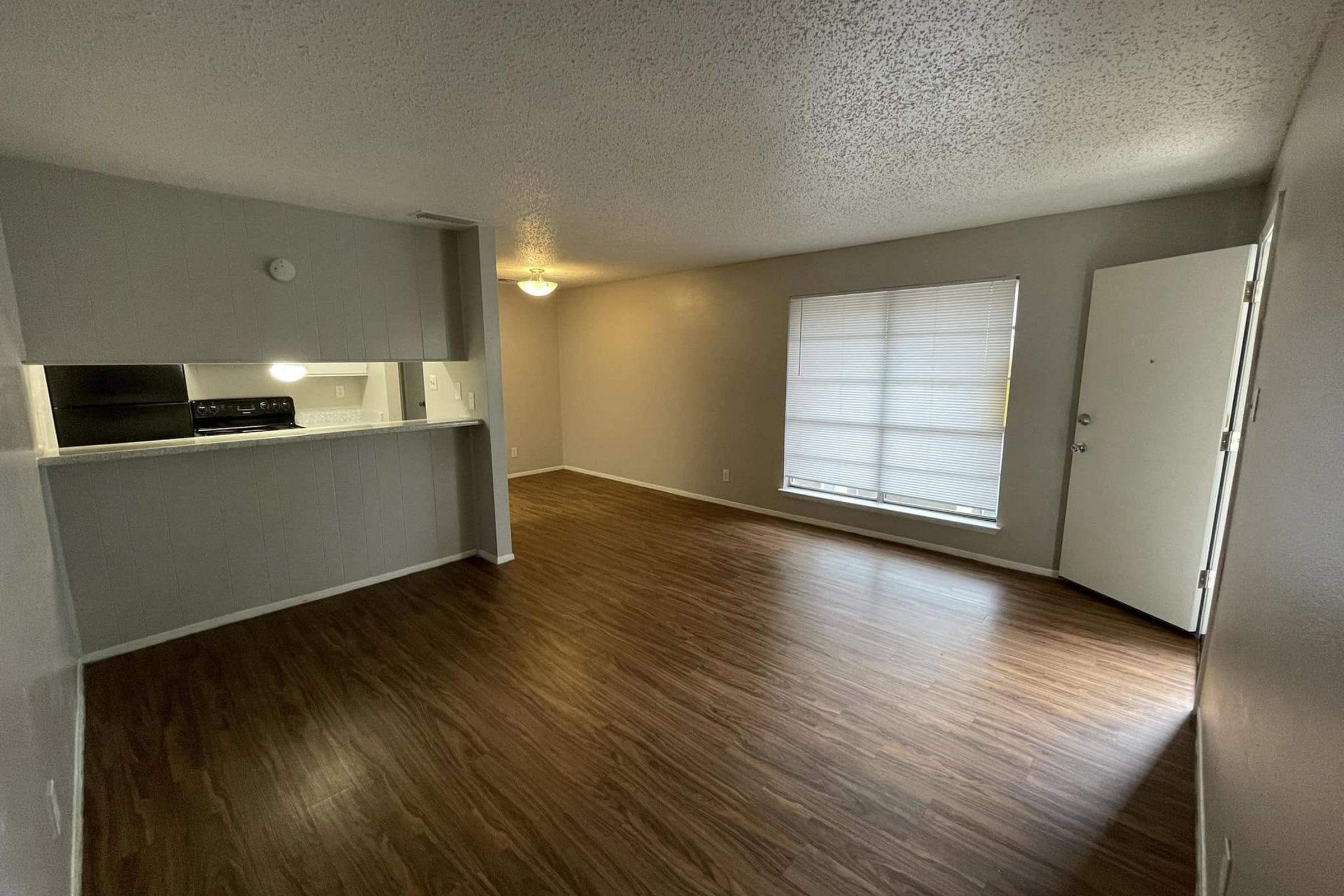 A spacious, empty living room with wood-like flooring, featuring a large window letting in natural light, a white wall with a door on the right, and a partial view of a kitchen area with a dark countertop and appliances in the background. The overall atmosphere is bright and inviting.
