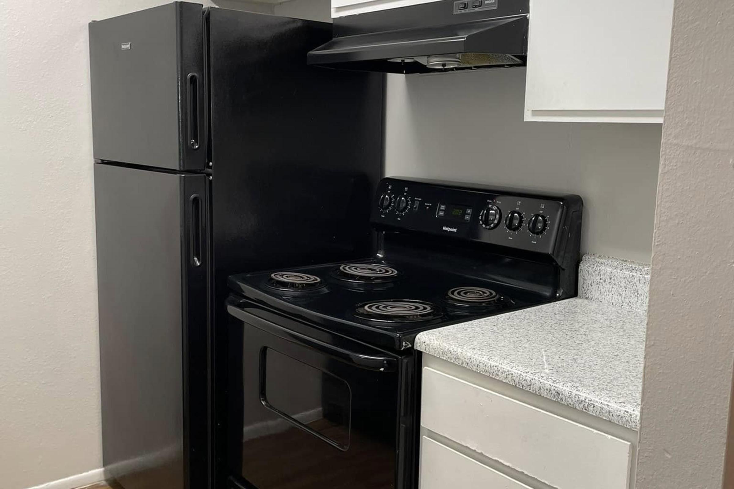 A compact kitchen featuring a black refrigerator, a black electric stove with four burners, and a light-colored countertop. The cabinetry above the stove is white, while the walls are a neutral tone, providing a simple and functional cooking space.