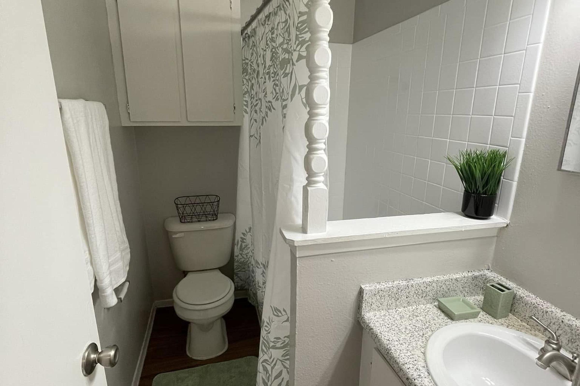A small bathroom featuring a toilet, a shower with a decorative curtain, a sink with a granite countertop, a potted plant, and neatly hung towels. The walls are painted a light gray, and there is a small storage area above the toilet. The overall design is clean and minimalist.