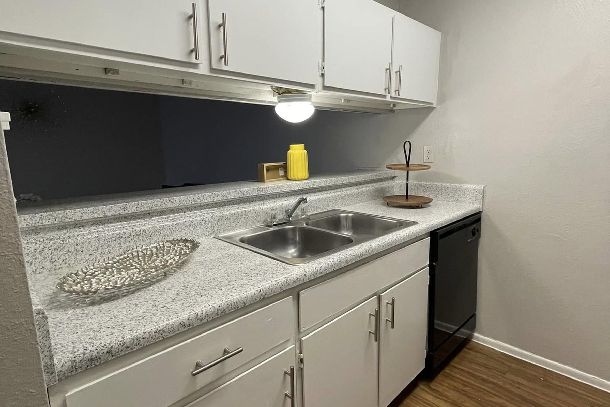A modern kitchen with a gray countertop featuring a double sink, a black dishwasher, and white cabinets. There's a circular wooden stand for displaying items and a decorative silver tray on the counter. A yellow container provides a pop of color against the neutral tones. Light illuminates the space.