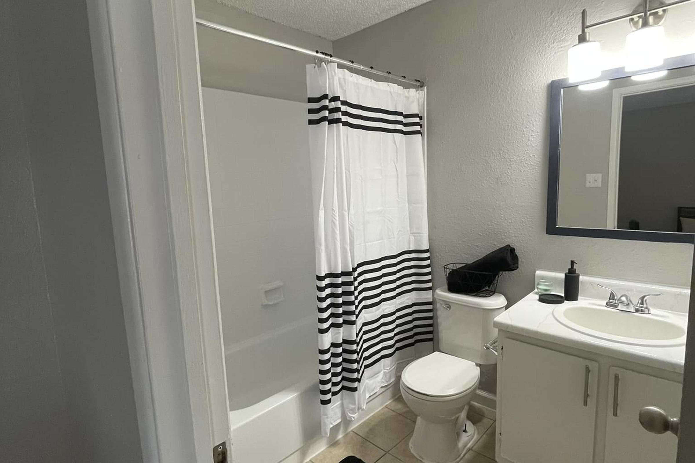 A well-lit bathroom featuring a bathtub with a shower curtain that has black and white stripes, a white toilet, and a sink with a mirror above it. The walls are painted a light gray, and the floor is tiled. A small black container is placed near the sink.