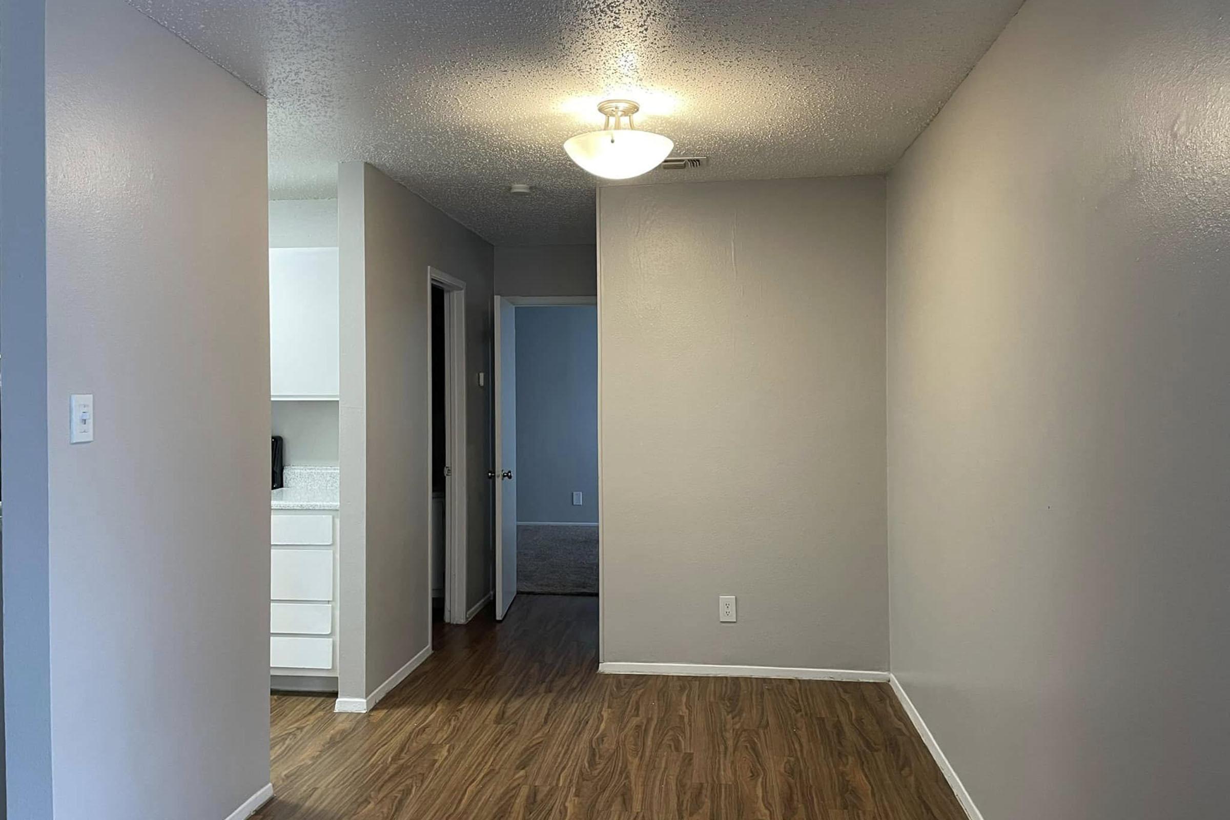 A well-lit interior hallway with a light fixture on the ceiling. The walls are painted in a light grey color, and the floor features wood-like laminate. On the left, there's a doorway leading to a kitchen area with white cabinets, while a carpeted room is visible through an open door on the right.