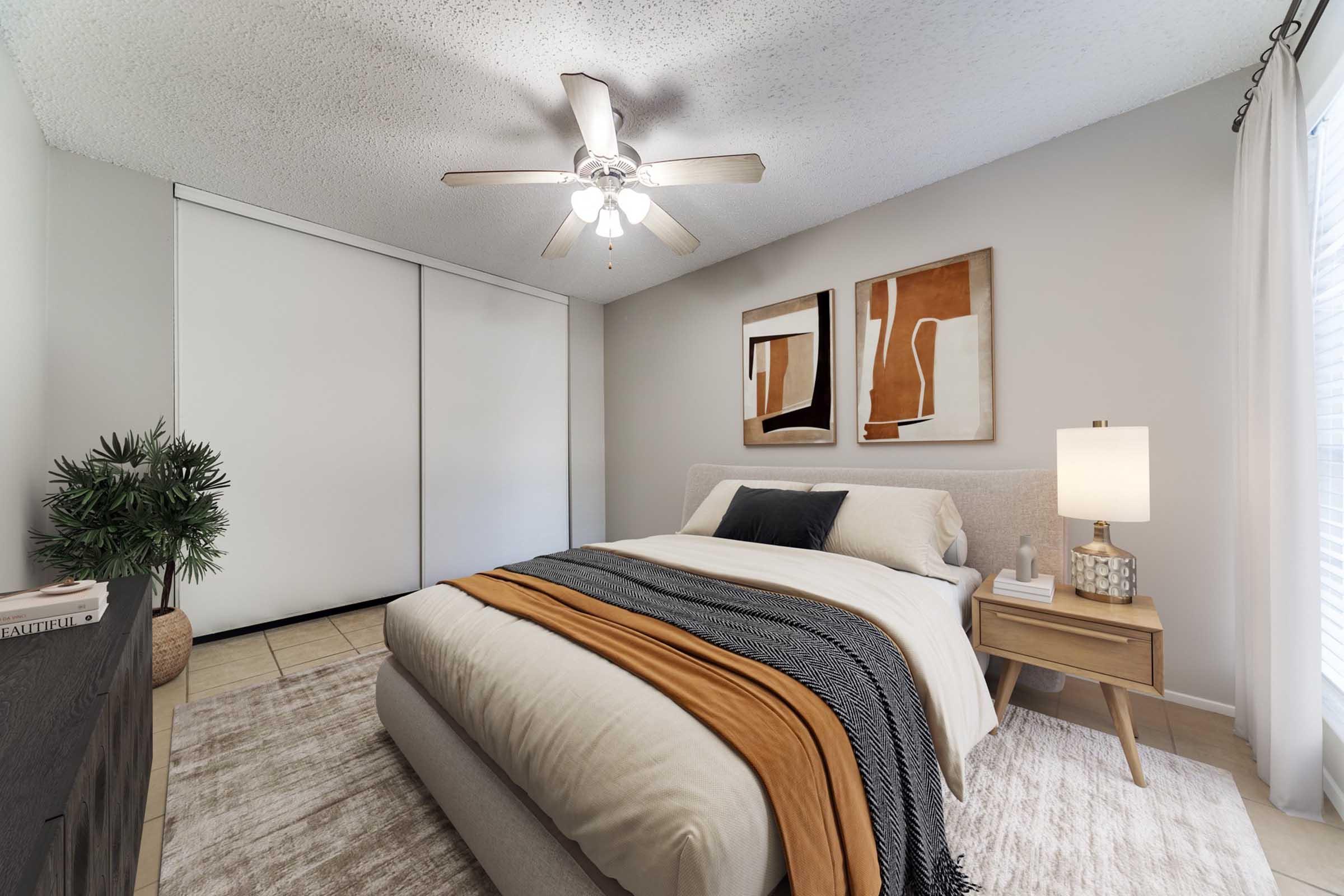 A cozy bedroom featuring a bed with neutral bedding and decorative throws. A bedside table with a lamp stands next to the bed. The room has light-colored walls, a ceiling fan, and two decorative framed artworks. Natural light filters through a window, and there's a potted plant accentuating the space.