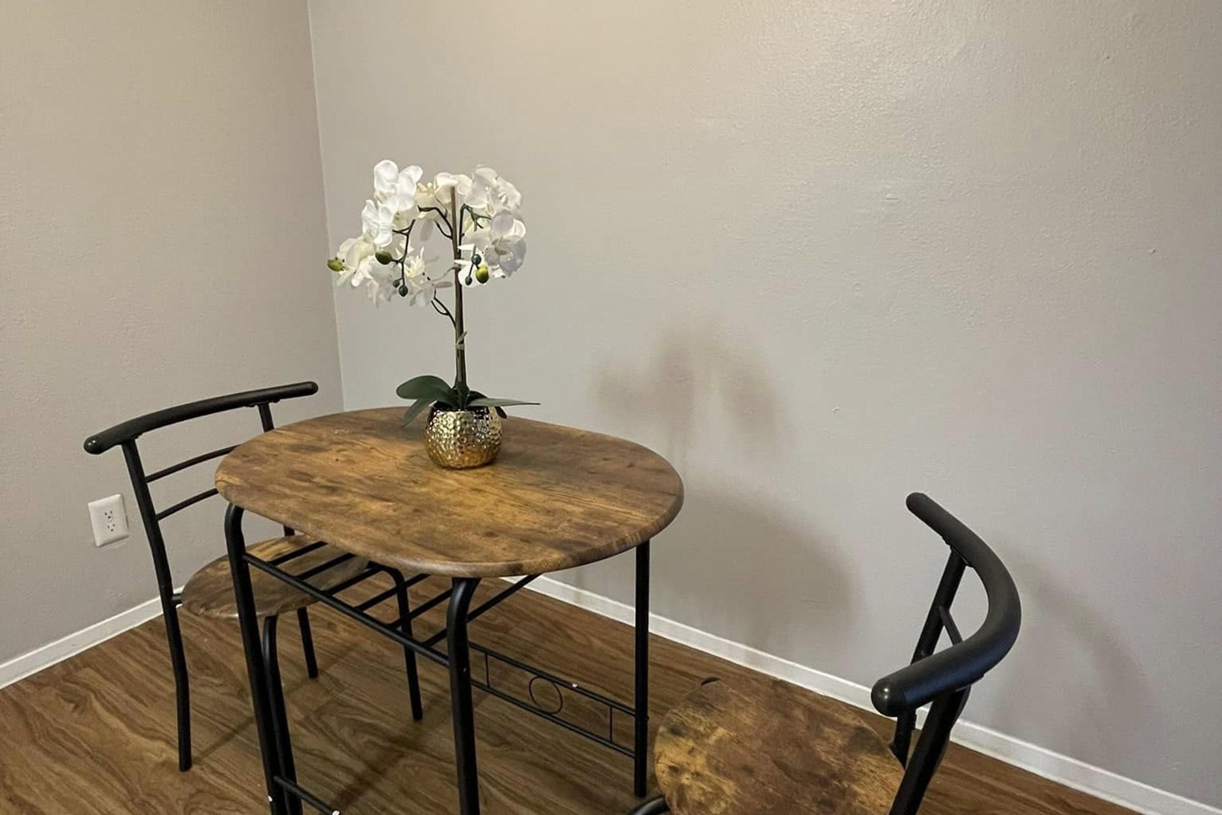 A small dining table made of wood with a textured surface, accompanied by two black metal chairs. A decorative vase with white orchids sits on the table, enhancing the simplicity of the setup. The walls are painted light gray, and the floor features wooden planks.