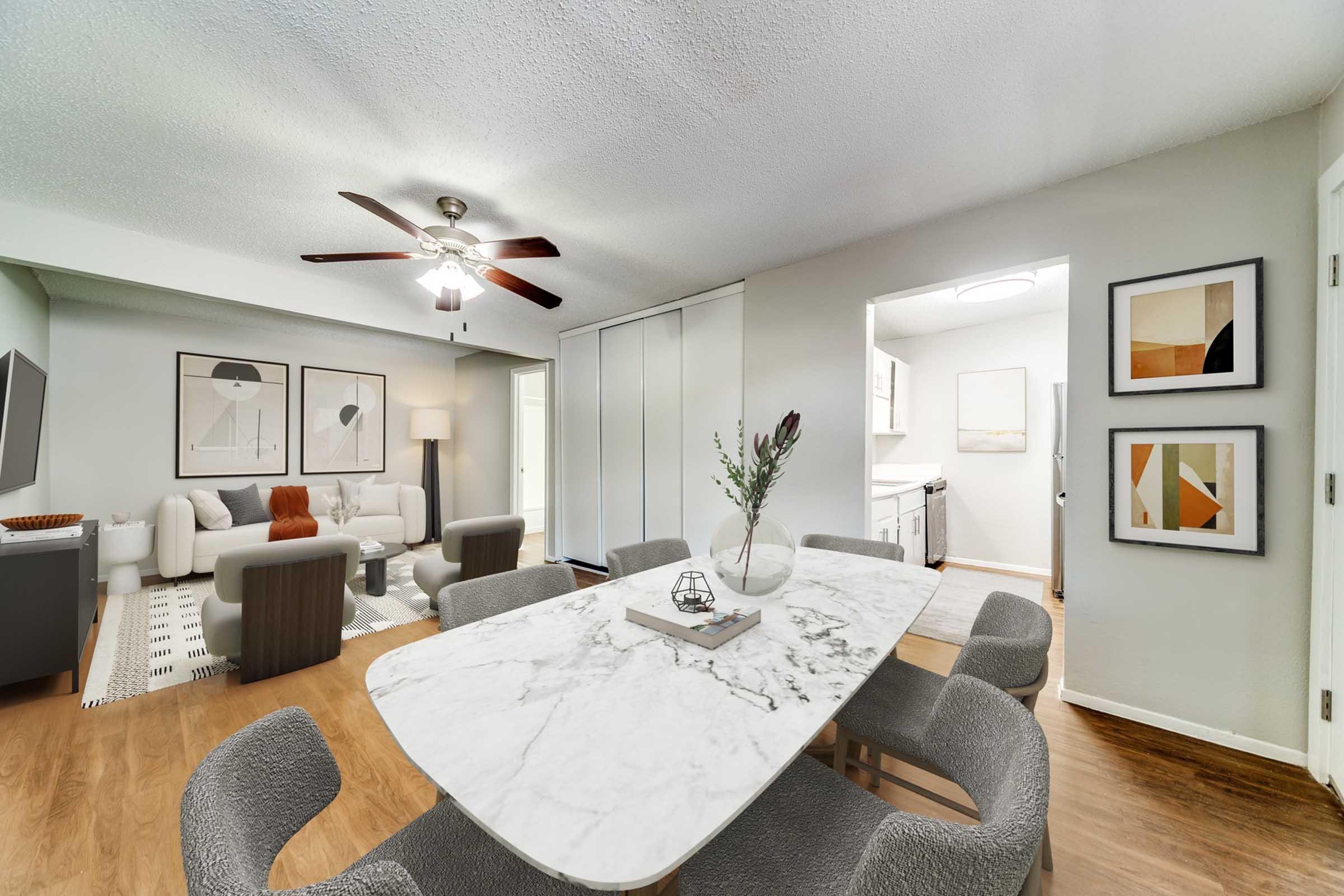 A spacious living and dining area featuring a marble dining table with grey chairs, a modern sofa set in the living space, and light-colored walls. A ceiling fan is visible, along with framed artwork on the wall and a view of a laundry area through sliding doors. The flooring is a warm wood tone.