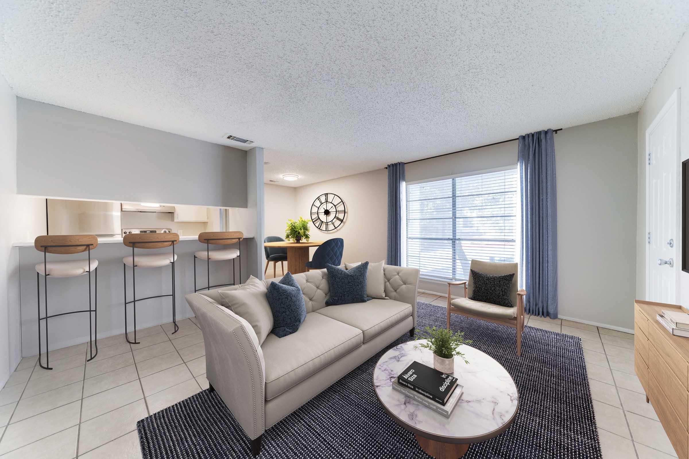 A cozy living room featuring a light-colored sofa with decorative pillows, a round coffee table, and modern decor. There are bar stools at a countertop that leads to a kitchen area. Natural light enters through a window dressed with curtains, creating a welcoming atmosphere.