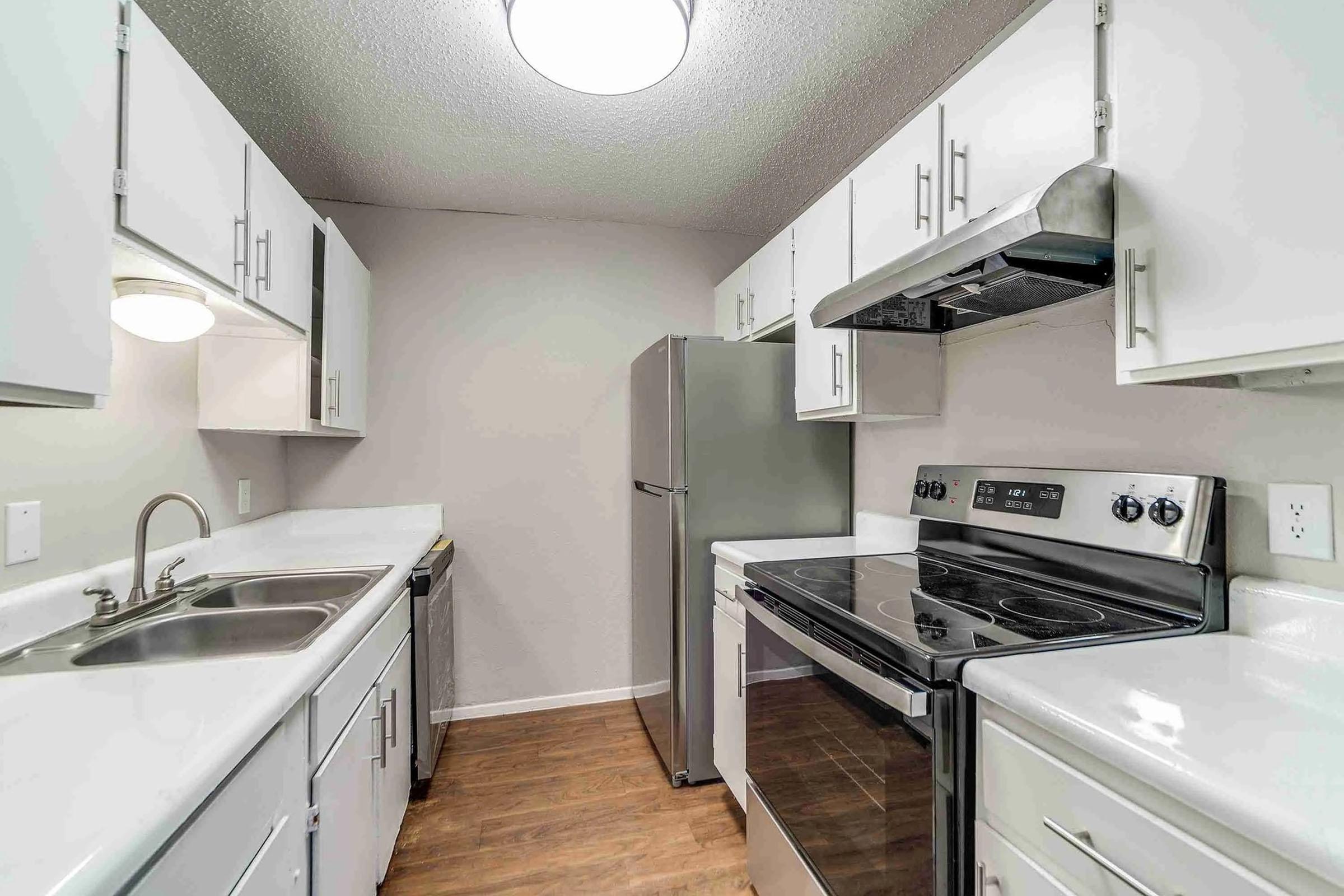 A modern kitchen with white cabinets, a stainless steel refrigerator, and a black electric stove. The countertops are light-colored, and the floors are wooden. Overhead lighting provides bright illumination, creating a clean and inviting atmosphere.