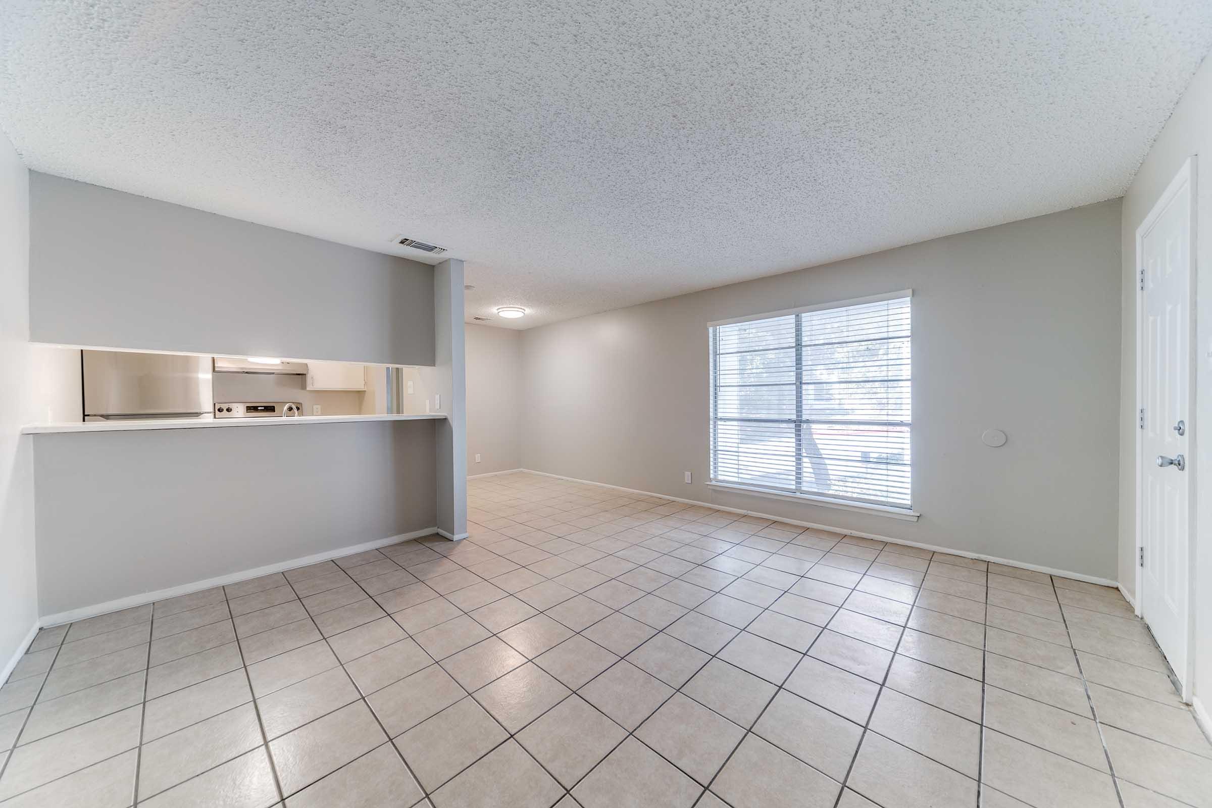 Spacious living area with tile flooring and neutral-colored walls. A large window allows natural light to fill the room. An open kitchen view is visible with modern appliances, and there is a doorway leading outside. The space is well-lit and inviting, suitable for various furniture arrangements.