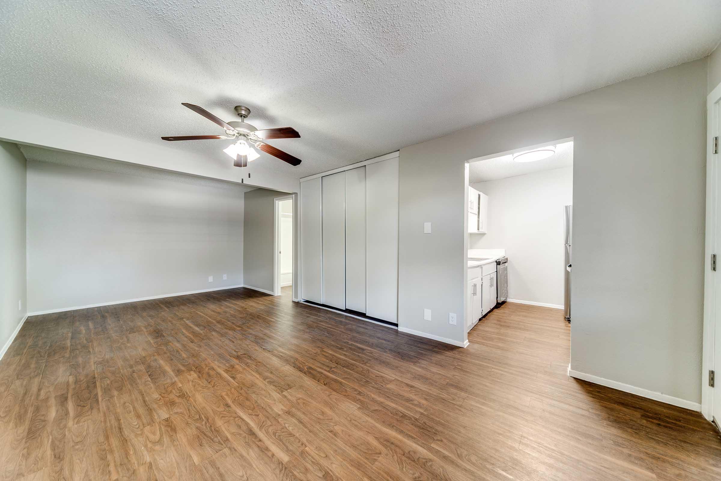 A spacious, modern living area with hardwood flooring and a ceiling fan. The room features neutral-colored walls and leads to a kitchen, visible on the right. Sliding doors are seen on the left, adding a functional element to the layout.
