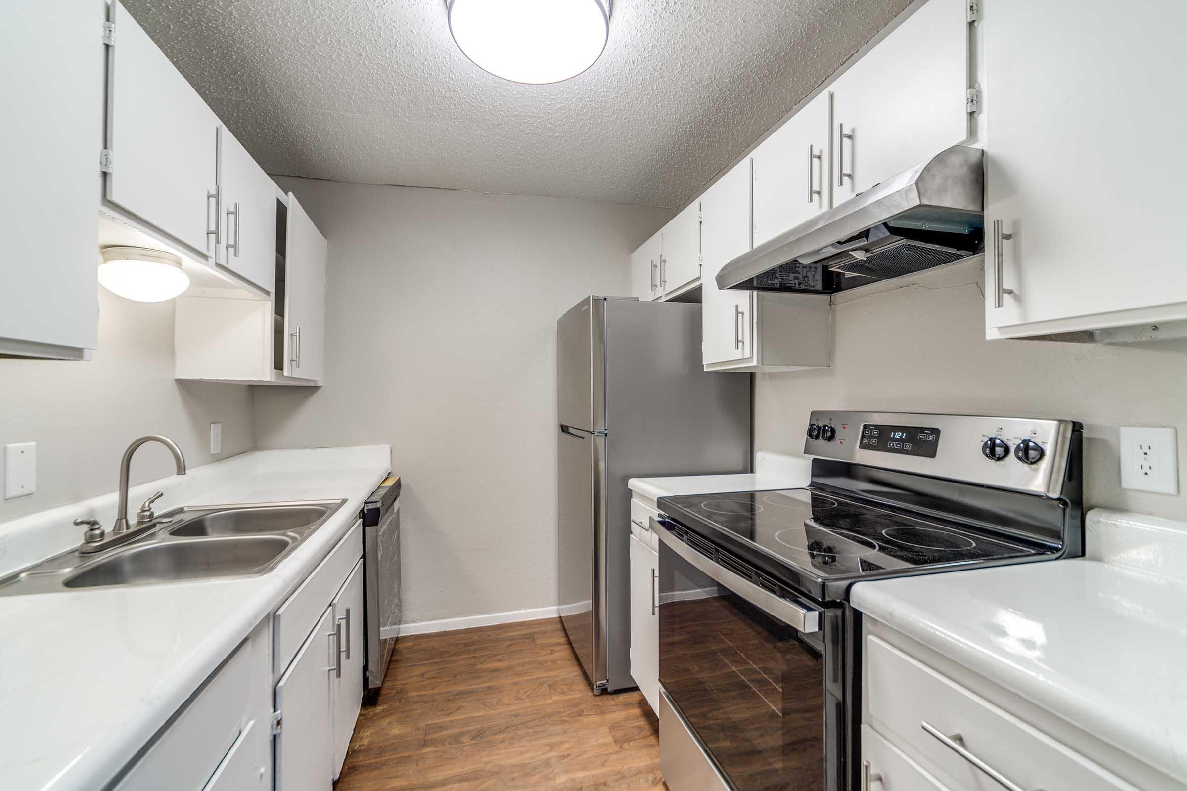 Modern kitchen featuring stainless steel appliances, including a refrigerator and stove. The space has white cabinetry with sleek handles, a double sink, and a large countertop. The flooring is wood-like, and the walls are painted a soft gray, creating a clean and inviting atmosphere.