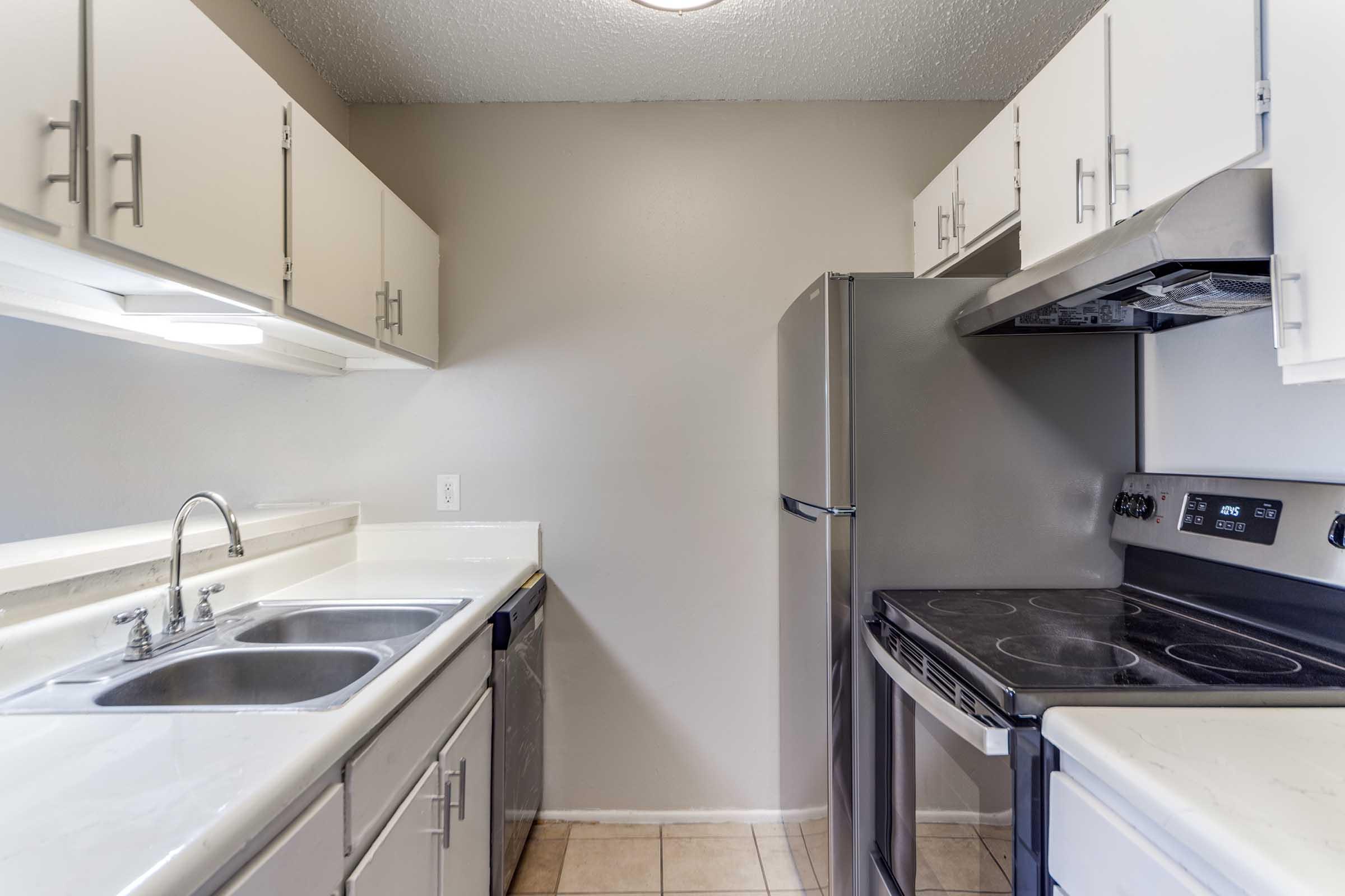 A modern kitchen featuring white cabinets, stainless steel appliances, including a refrigerator and an oven/stove combo, and a double sink. The walls are painted a light grey, and the floor is tiled. The layout is functional, with ample storage in the cabinetry.