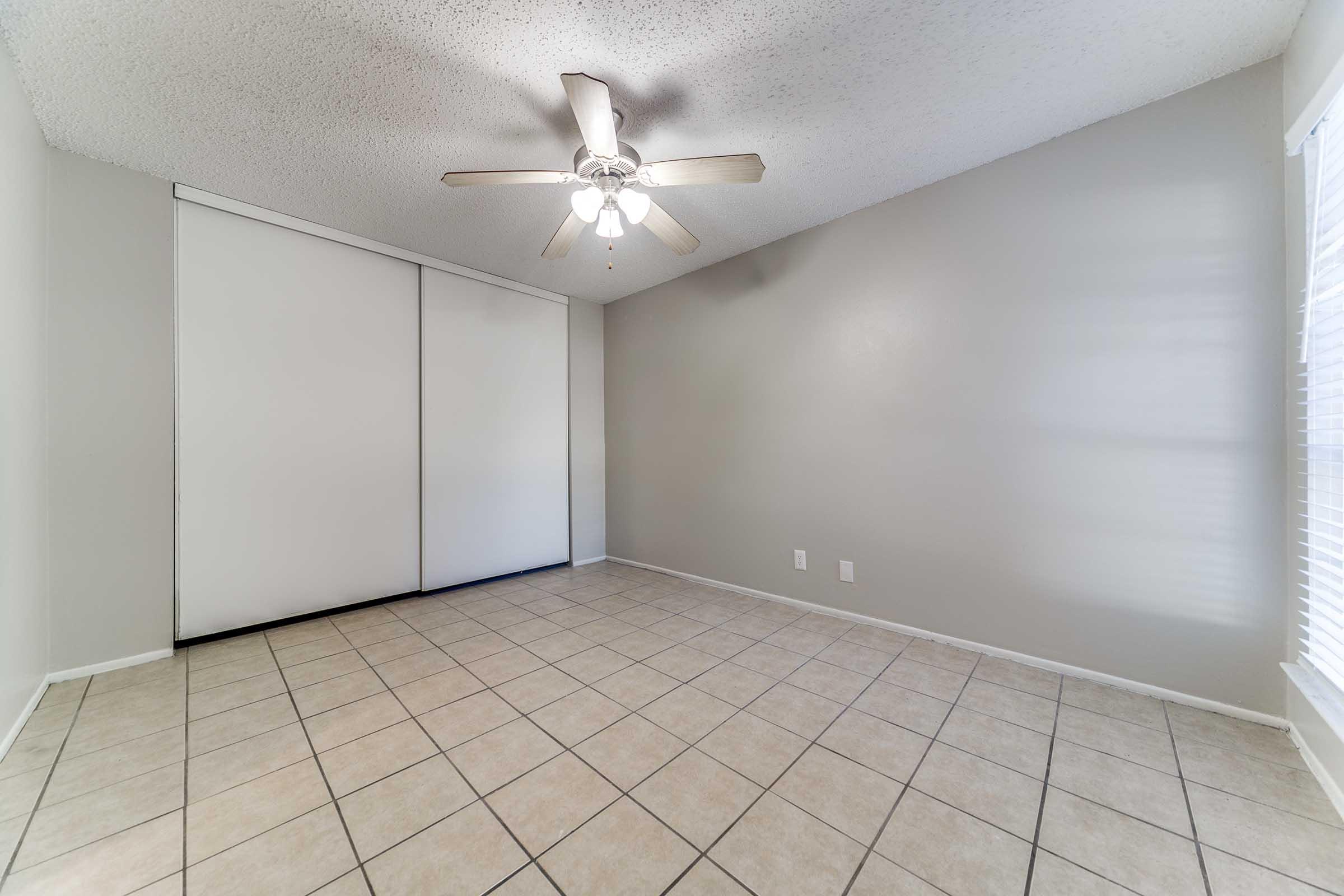 Empty room with a ceiling fan, light fixture, and beige walls. The floor is tiled in a light color. There are large sliding closet doors on one wall and a window with white blinds on another wall, allowing natural light to enter the space.