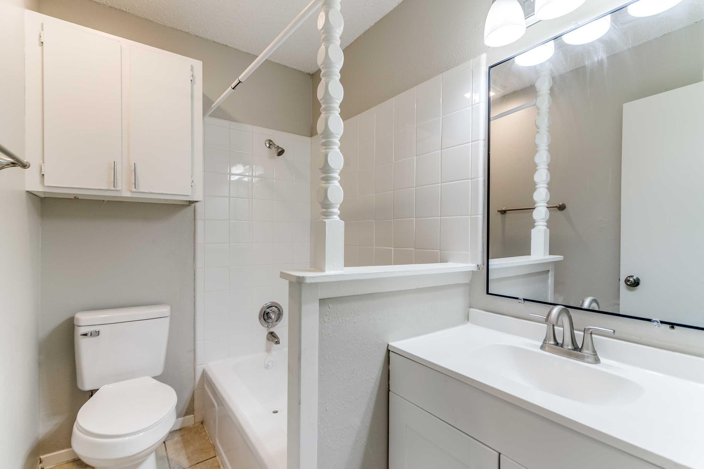 A clean bathroom featuring a white toilet, a bathtub with a shower, a modern sink with a mirror above, and white cabinetry. The walls are painted in a neutral color, and there is good lighting from the overhead fixture. The overall design is functional and contemporary.