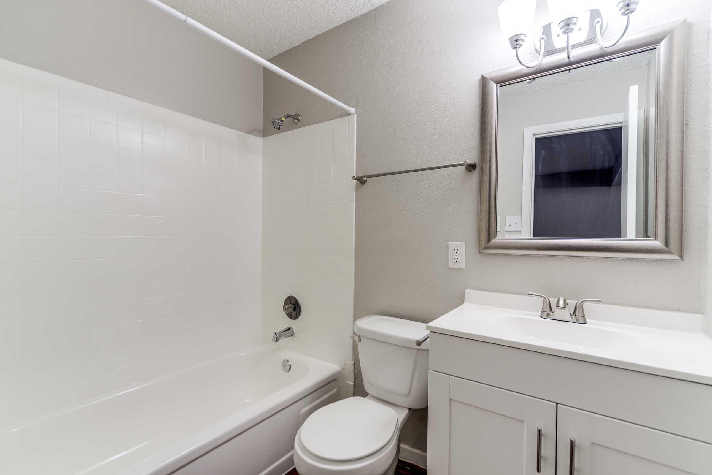A modern bathroom featuring a white bathtub with a shower, a sleek vanity with a sink and mirror, and a toilet. The walls are painted in a light gray color, and there is a light fixture above the mirror. The space is bright and clean, conveying a fresh and minimalist aesthetic.