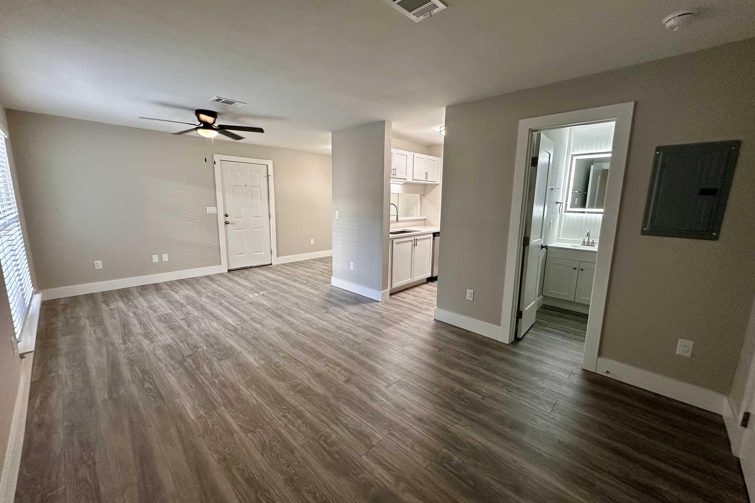 A spacious interior of an apartment features a living area with a ceiling fan, light-colored walls, and laminate wood flooring. On one side, there’s a doorway leading to a small bathroom with a sink and mirror. The kitchen area is partially visible with cabinets in the background. Natural light comes through the windows.